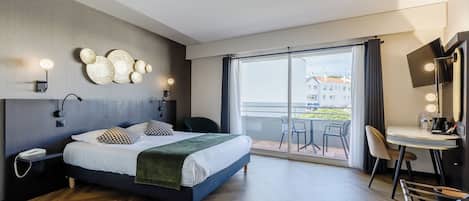 Ein modernes Hotelzimmer mit einem großen Bett, einem Schreibtisch mit Stuhl, einem Balkon mit Blick auf Gebäude und Wandleuchten.