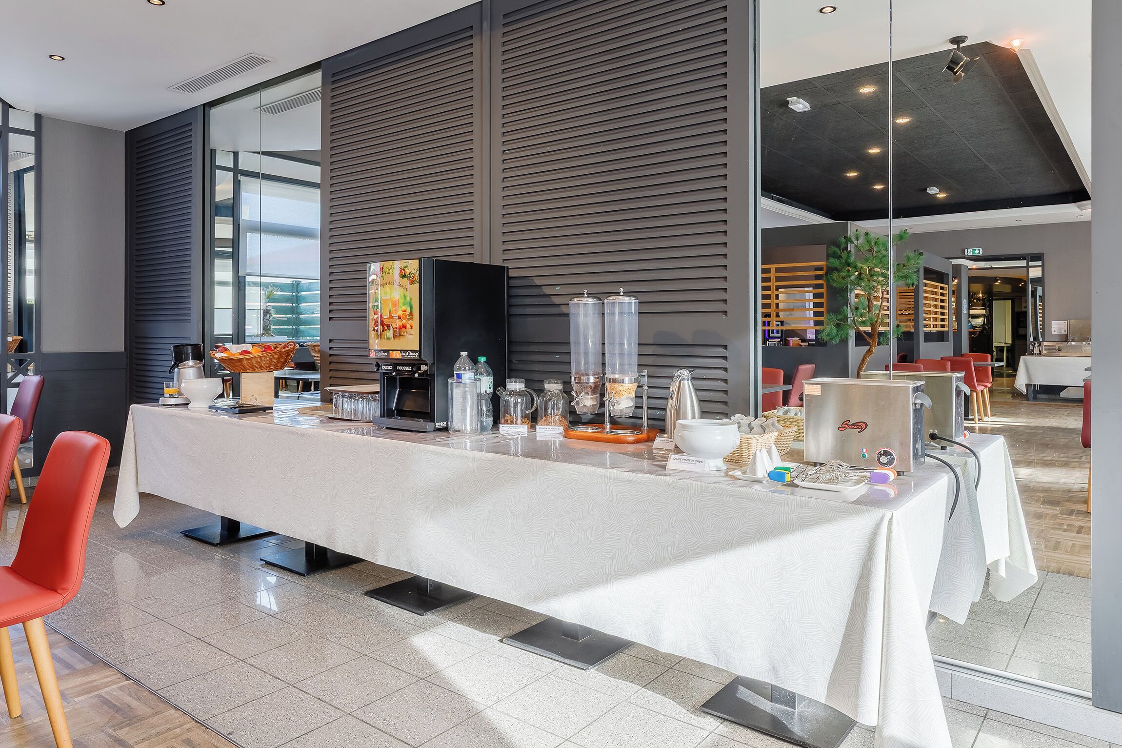 Una habitación de hotel moderna con una larga mesa de comedor, sillas rojas, una cafetera y un amplio ventanal con vistas a la ciudad.