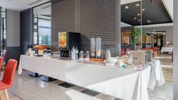 Moderni hotellihuone, jossa on pitkä ruokapöytä, punaiset tuolit, kahvinkeitin ja suuri ikkuna, josta avautuu näkymä kaupunkiin.