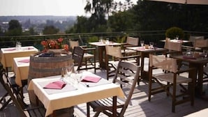 Un área de comedor al aire libre con mesas y sillas, algunas con sombrillas, frente a un fondo de árboles y una vista distante.