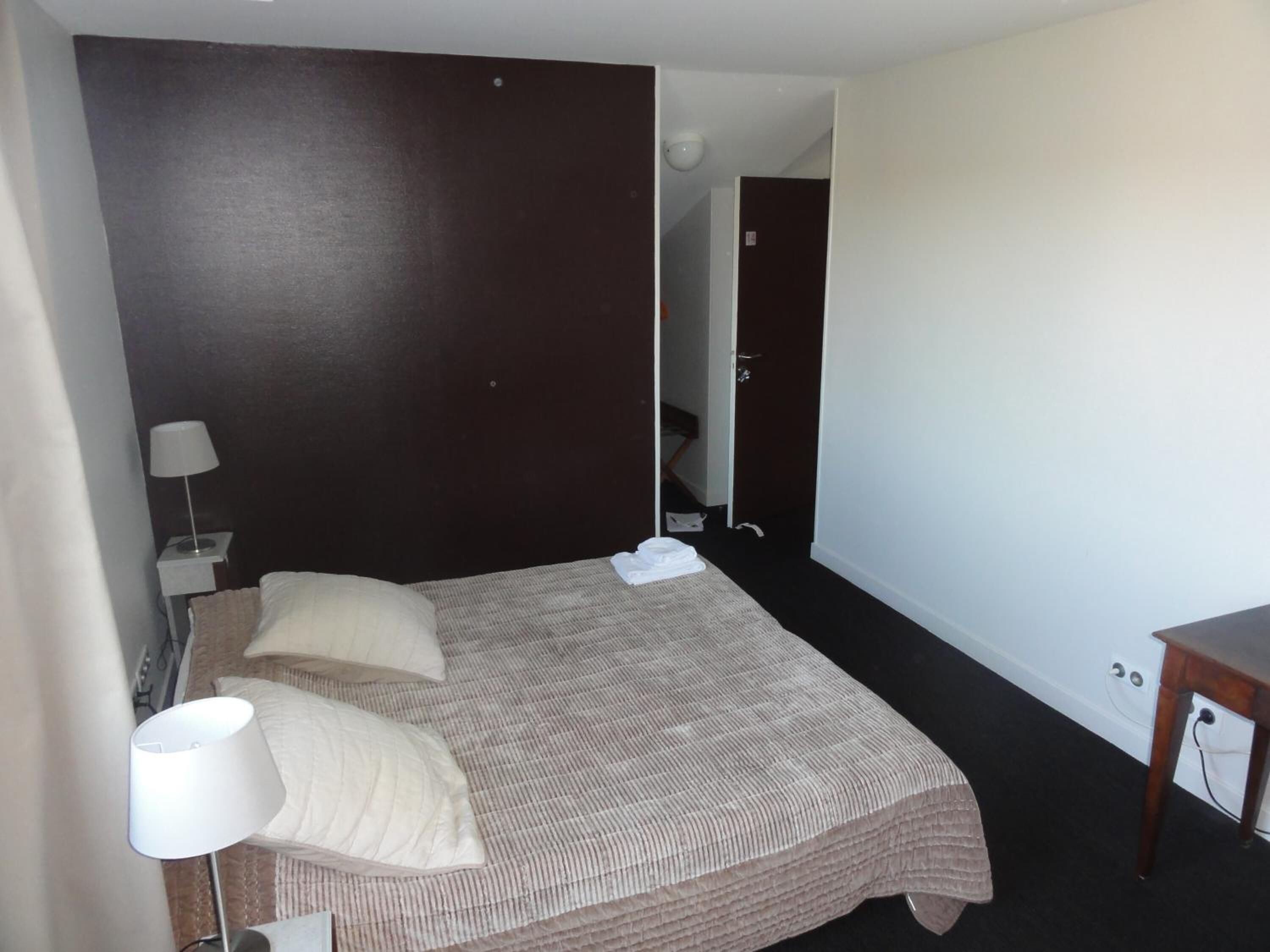 Quarto de hotel com uma cama grande, duas almofadas, um abajur e uma mesinha.