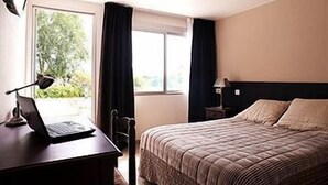 Una habitación de hotel con una cama, un escritorio con una computadora portátil, una silla, una ventana con cortinas y una mesita de noche con una lámpara.