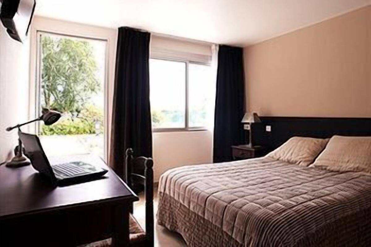 Une chambre d’hôtel comprenant un lit, un bureau avec un ordinateur portable, une chaise, une fenêtre avec des rideaux et une table de chevet avec une lampe.