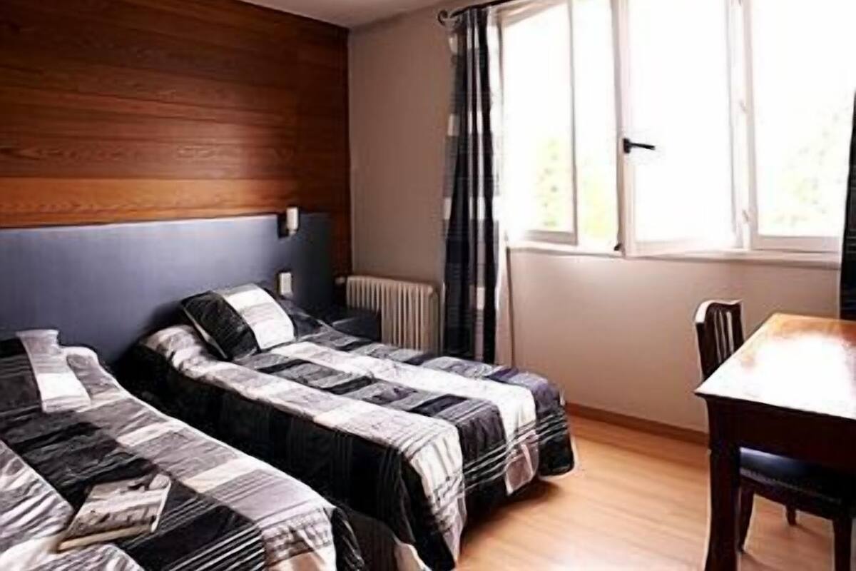Une chambre avec deux lits, une tête de lit en bois, une fenêtre avec des rideaux et une table en bois avec une chaise.