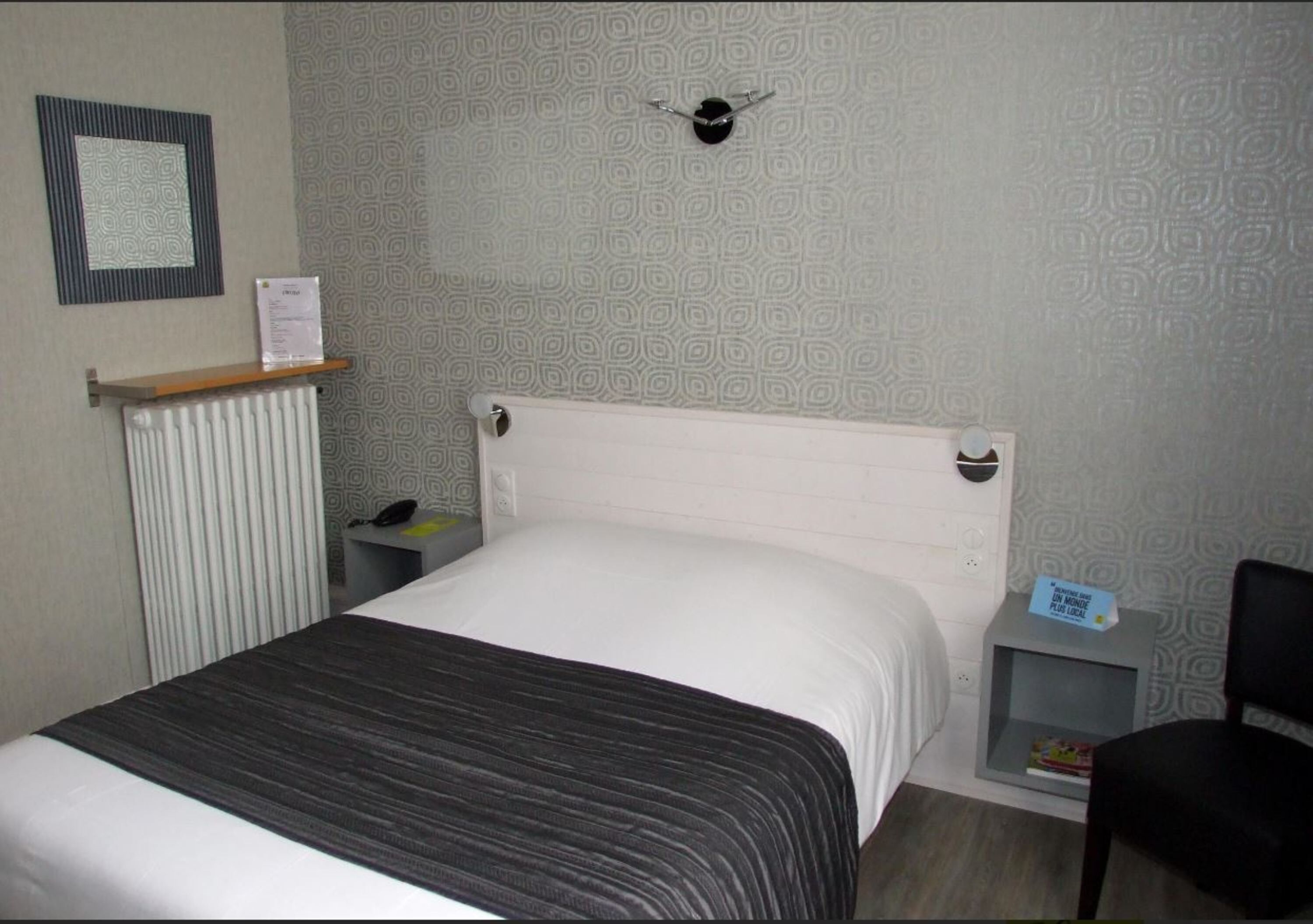 Habitació d'hotel amb un llit, una taula de nit amb una làmpada, un radiador, una petita estant i un rellotge penjat a la paret.
