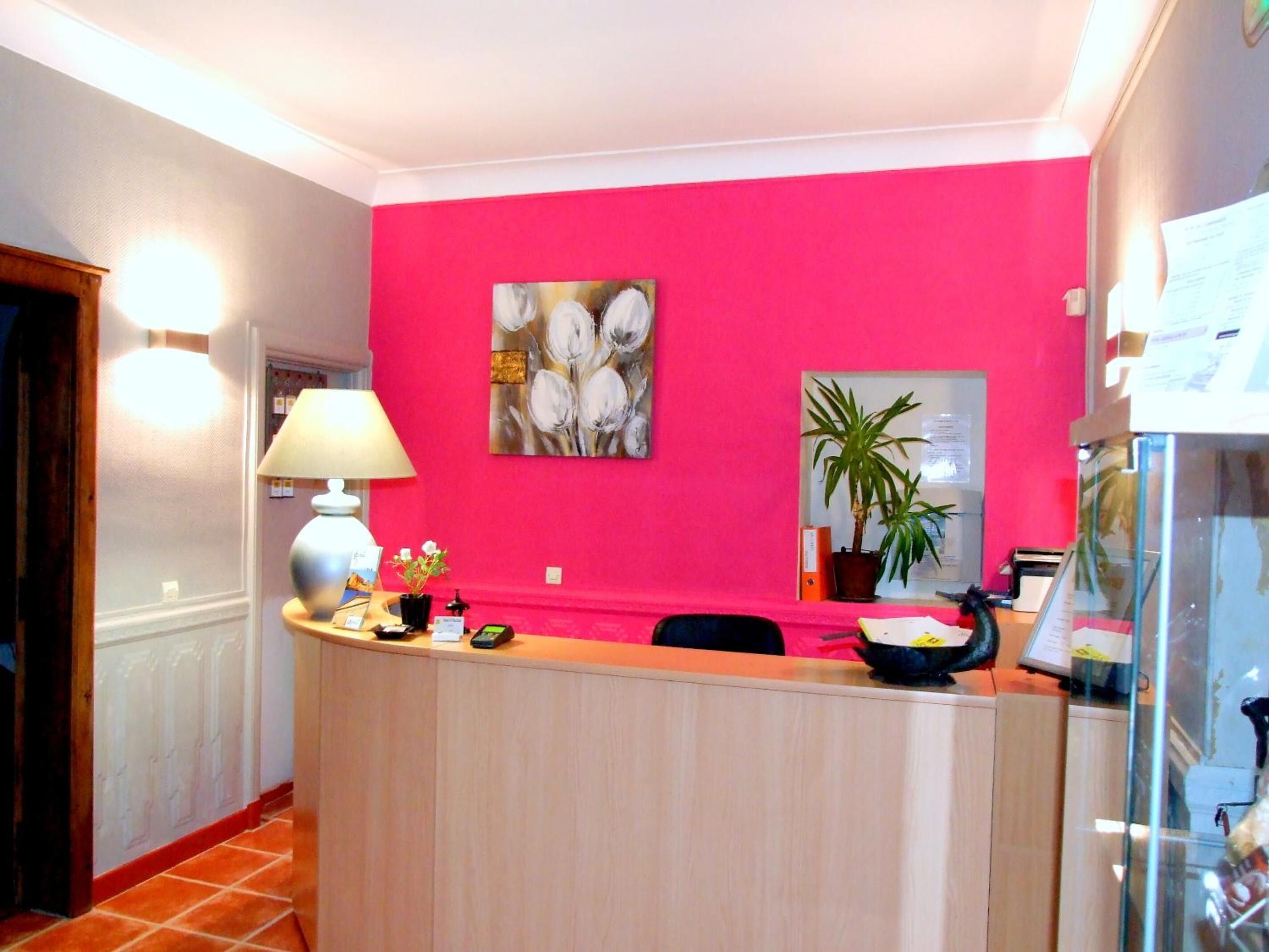 A reception area with a pink wall, a painting, a desk, a lamp, a potted plant, and a glass display case.