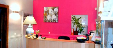 A reception area with a pink wall, a painting, a desk, a lamp, a potted plant, and a glass display case.