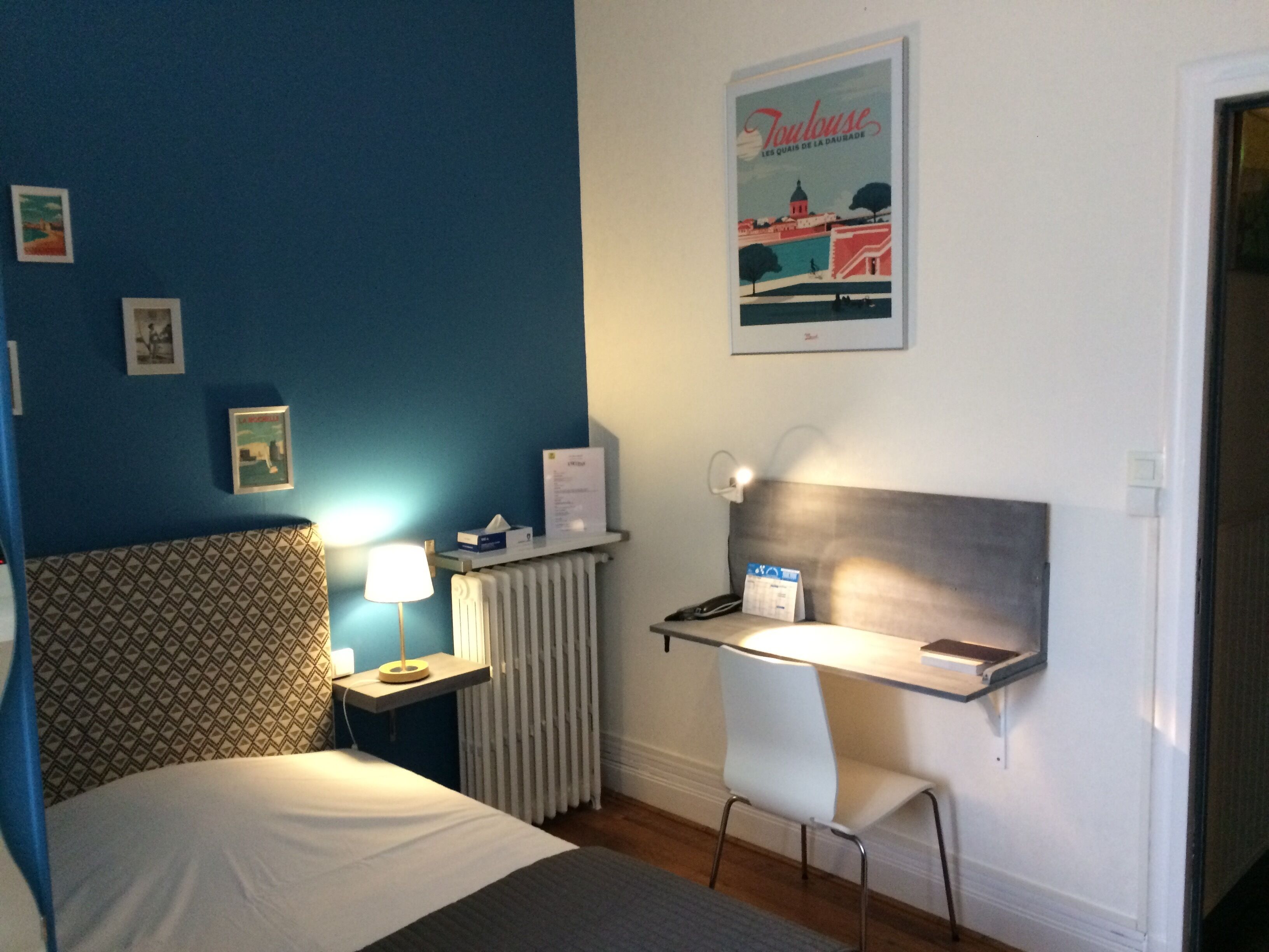 A hotel room with a bed, a desk, a chair, a lamp, a radiator, and a poster on the wall.