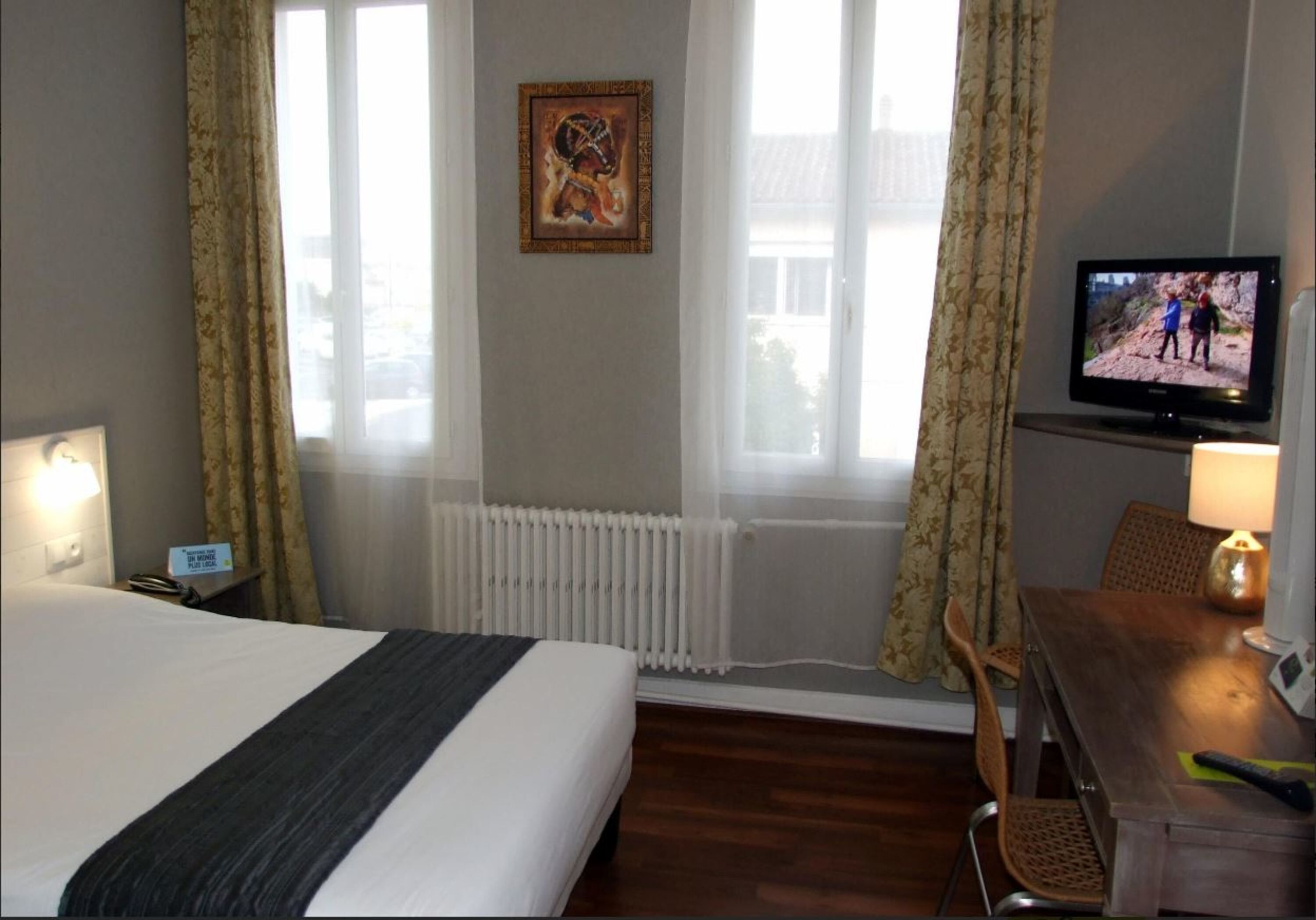A hotel room with a bed, a television, a desk with a lamp, and a window with curtains.