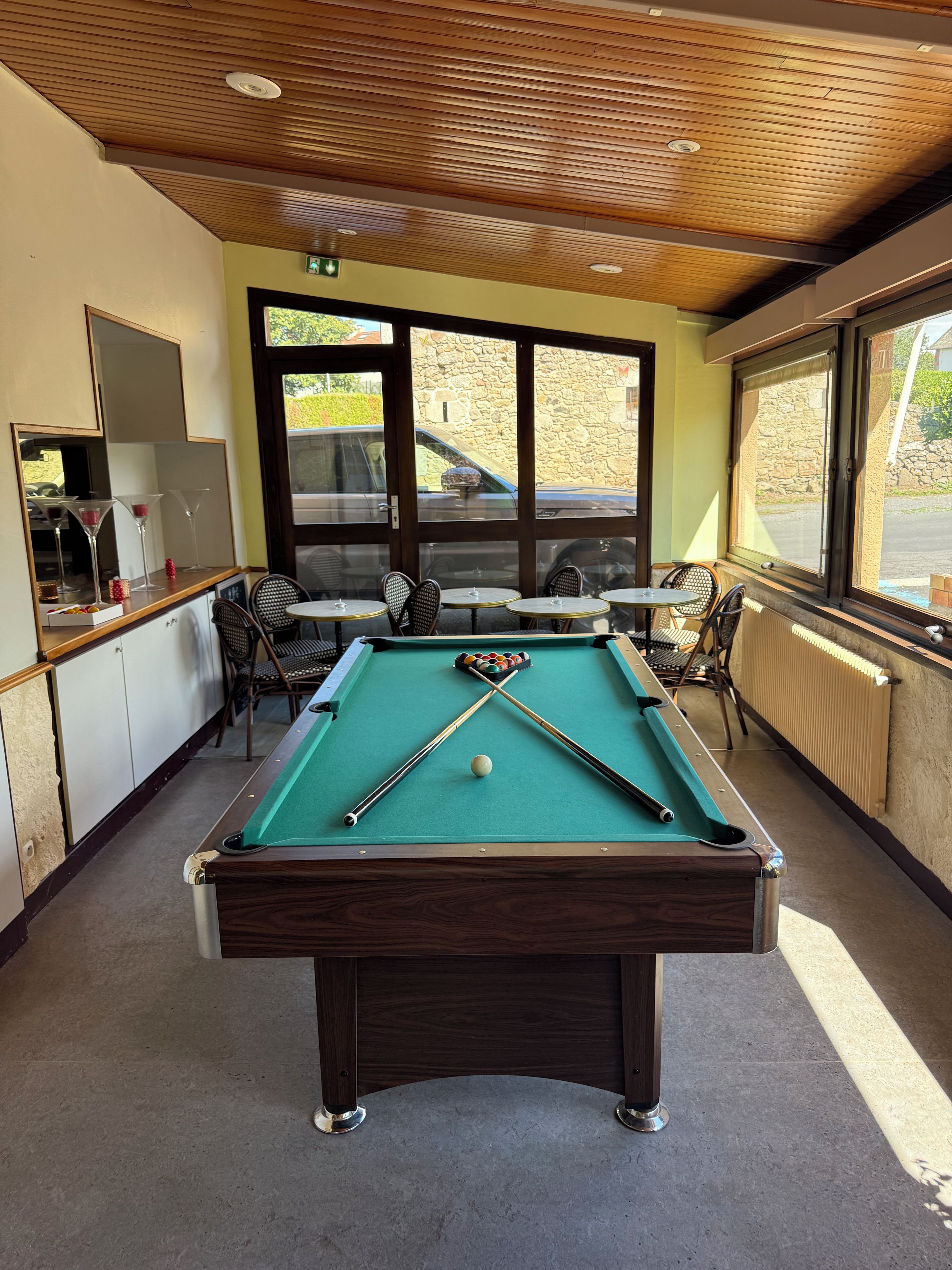 A pool table with cues and balls, surrounded by chairs and a table, with windows and a view of the outside.