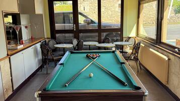 A pool table with cues and balls, surrounded by chairs and a table, with windows and a view of the outside.