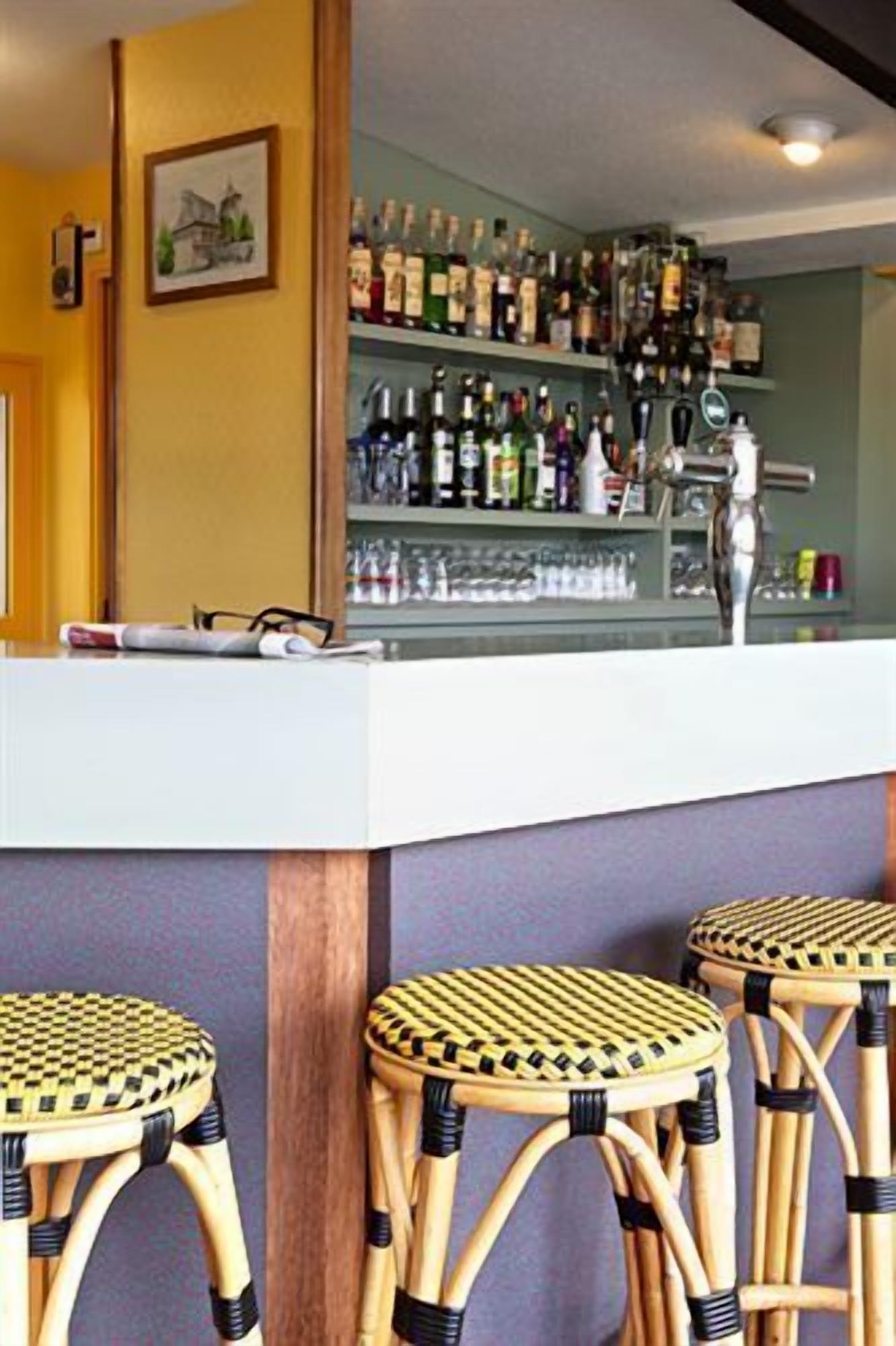 Una zona de bar con mostrador, estantes llenos de botellas y tres taburetes de mimbre.
