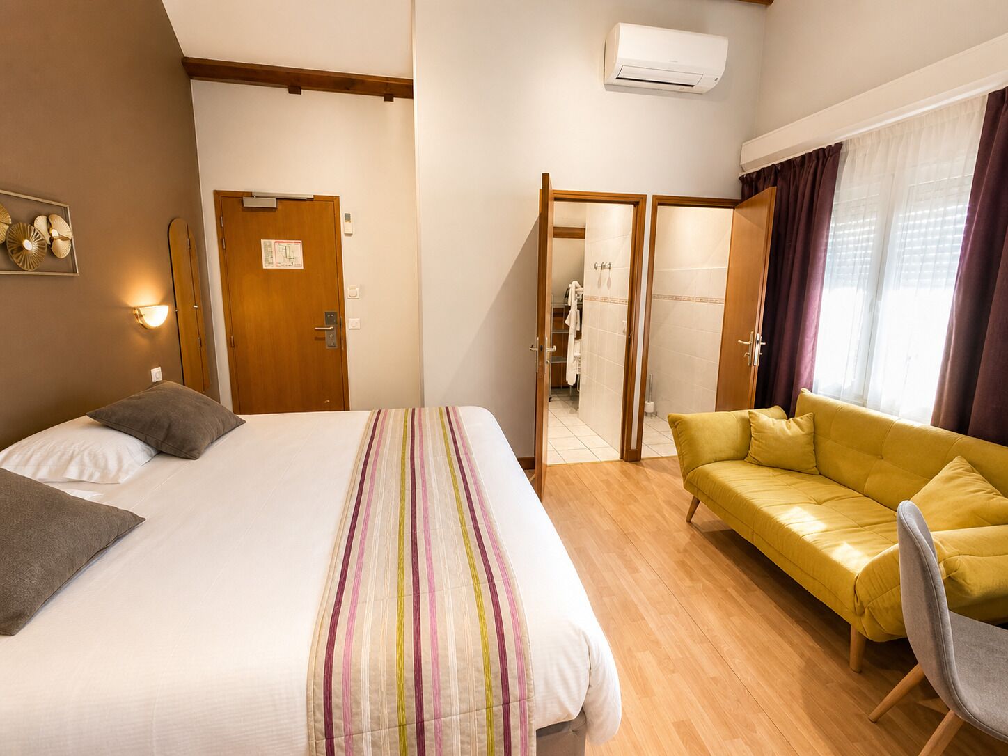 Habitación de hotel con cama, sofá, sillón, unidad de aire acondicionado y puerta que da a un baño.