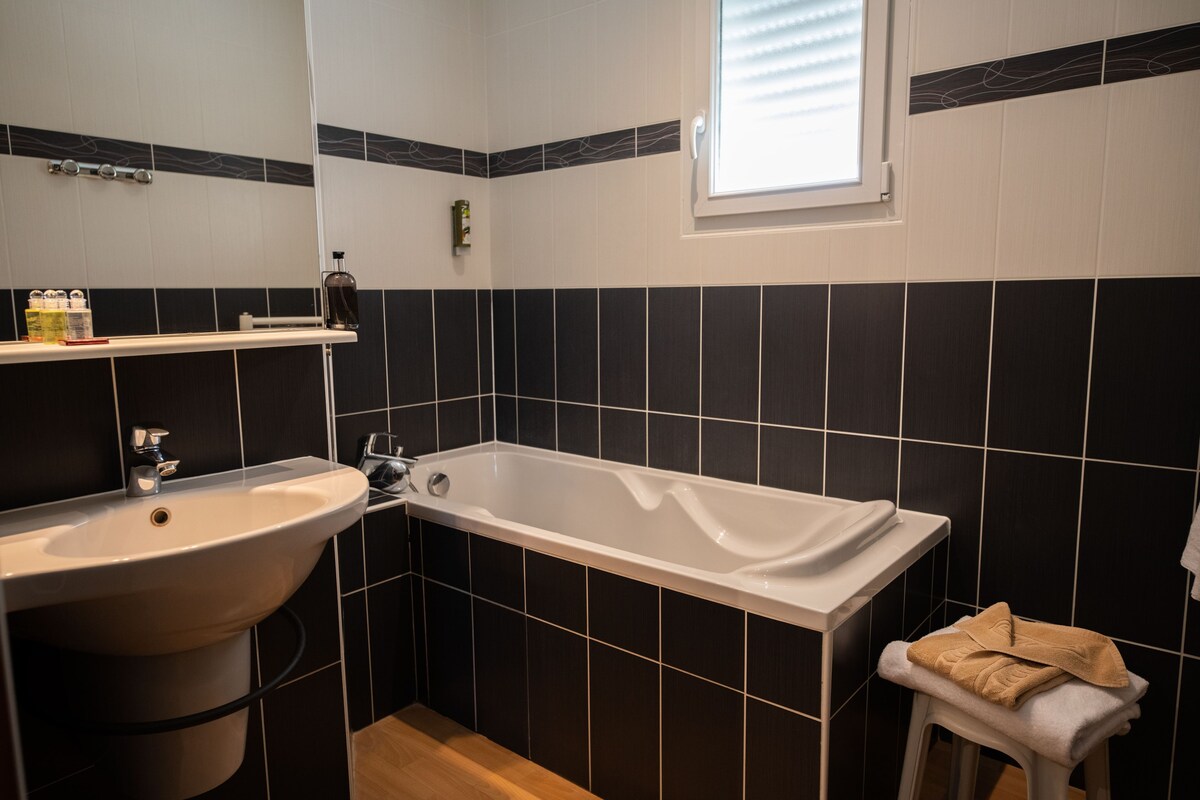 A bathroom with a bathtub, a sink, a window, and a towel on a stool.
