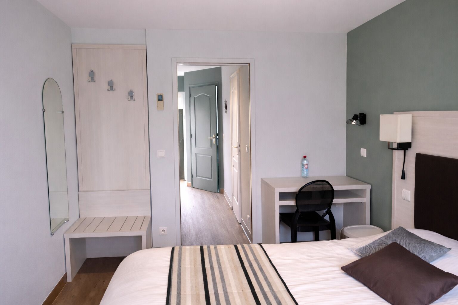 A hotel room with a bed, a desk, a chair, a mirror, and a door leading to another room.