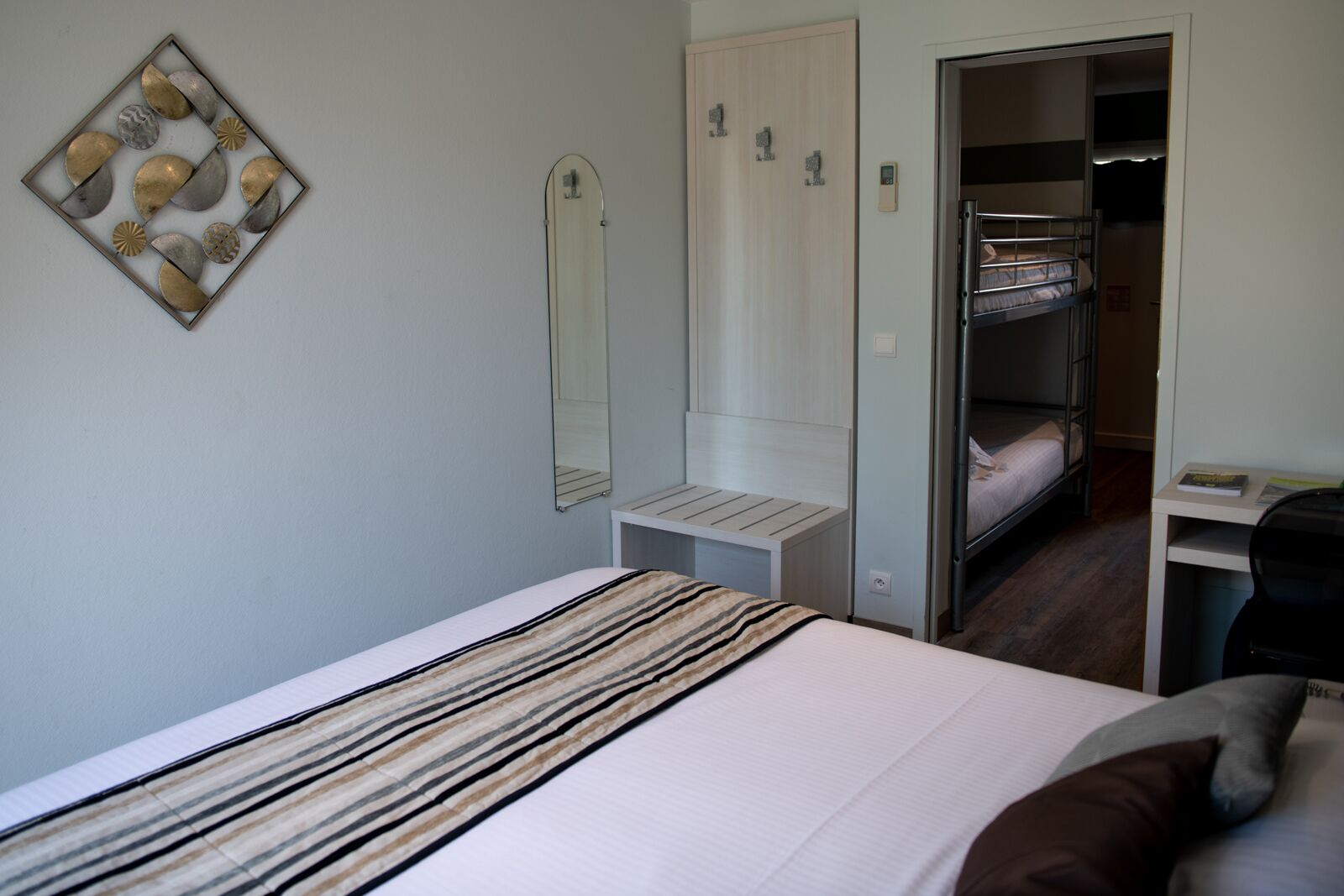 A bedroom with a bed, a bedside table, a mirror, a wardrobe, and a bunk bed in the background.