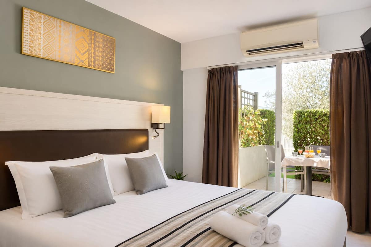 A hotel room with a bed, a headboard, a wall-mounted lamp, a window with curtains, and an air conditioning unit.