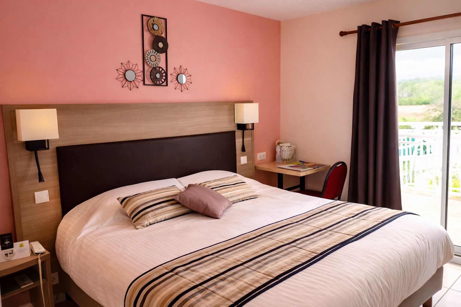Een hotelkamer met een groot bed, nachtlampjes, een klein tafeltje met een stoel, en uitzicht op een balkon.