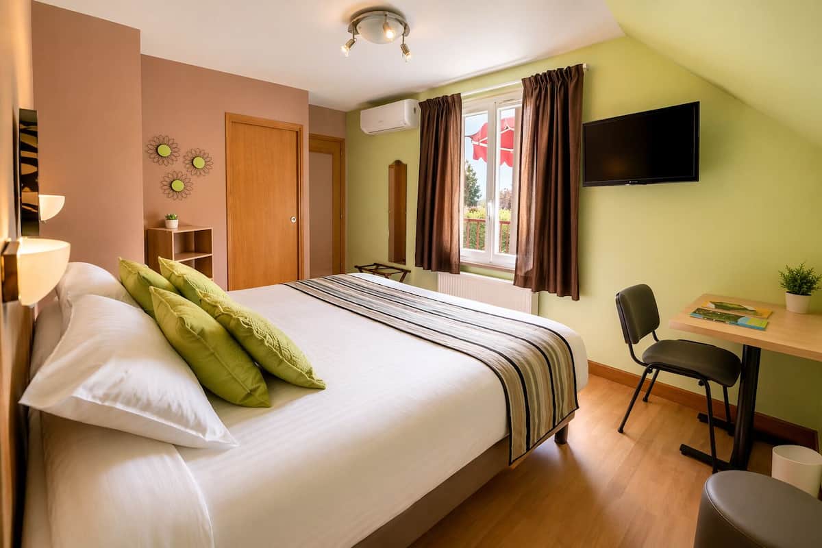 A bedroom with a bed, a desk, a chair, a TV, and a window with curtains.