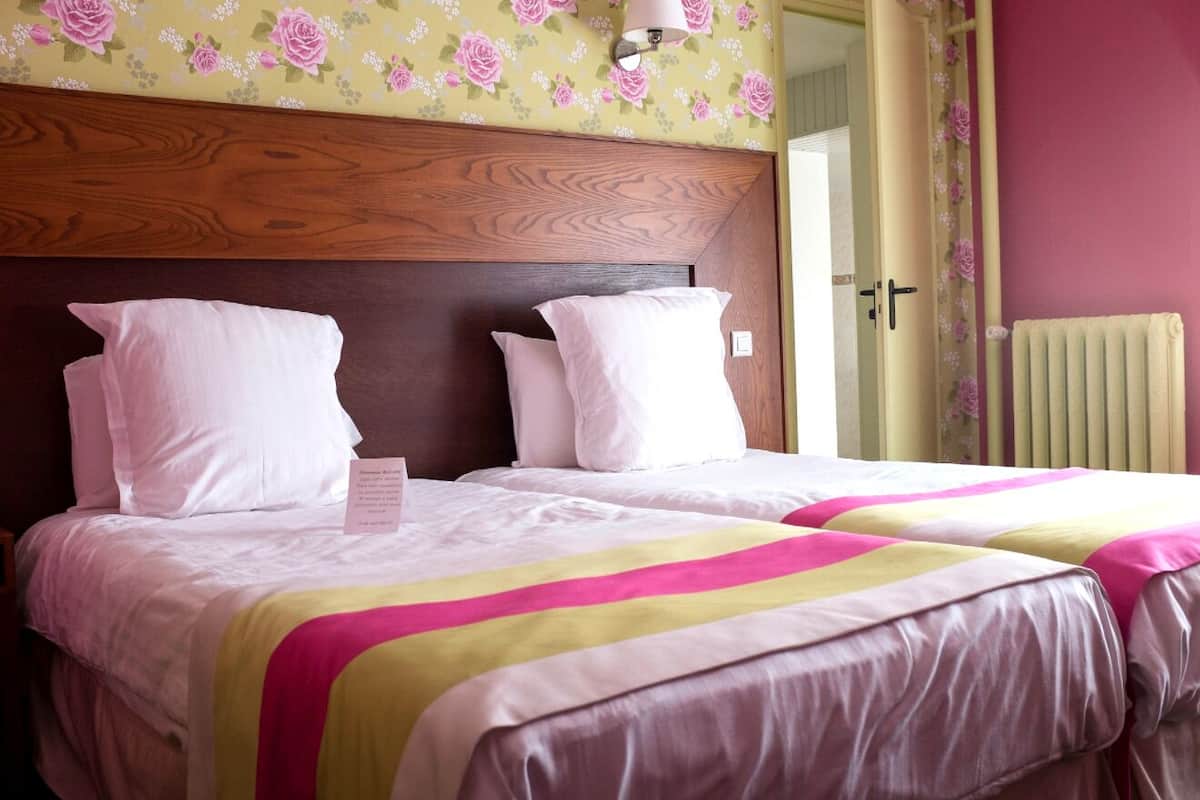 Una habitación de hotel con cabecera de madera, dos camas con ropa blanca y mantas a rayas rosas y amarillas, papel tapiz floral y un radiador en la pared.