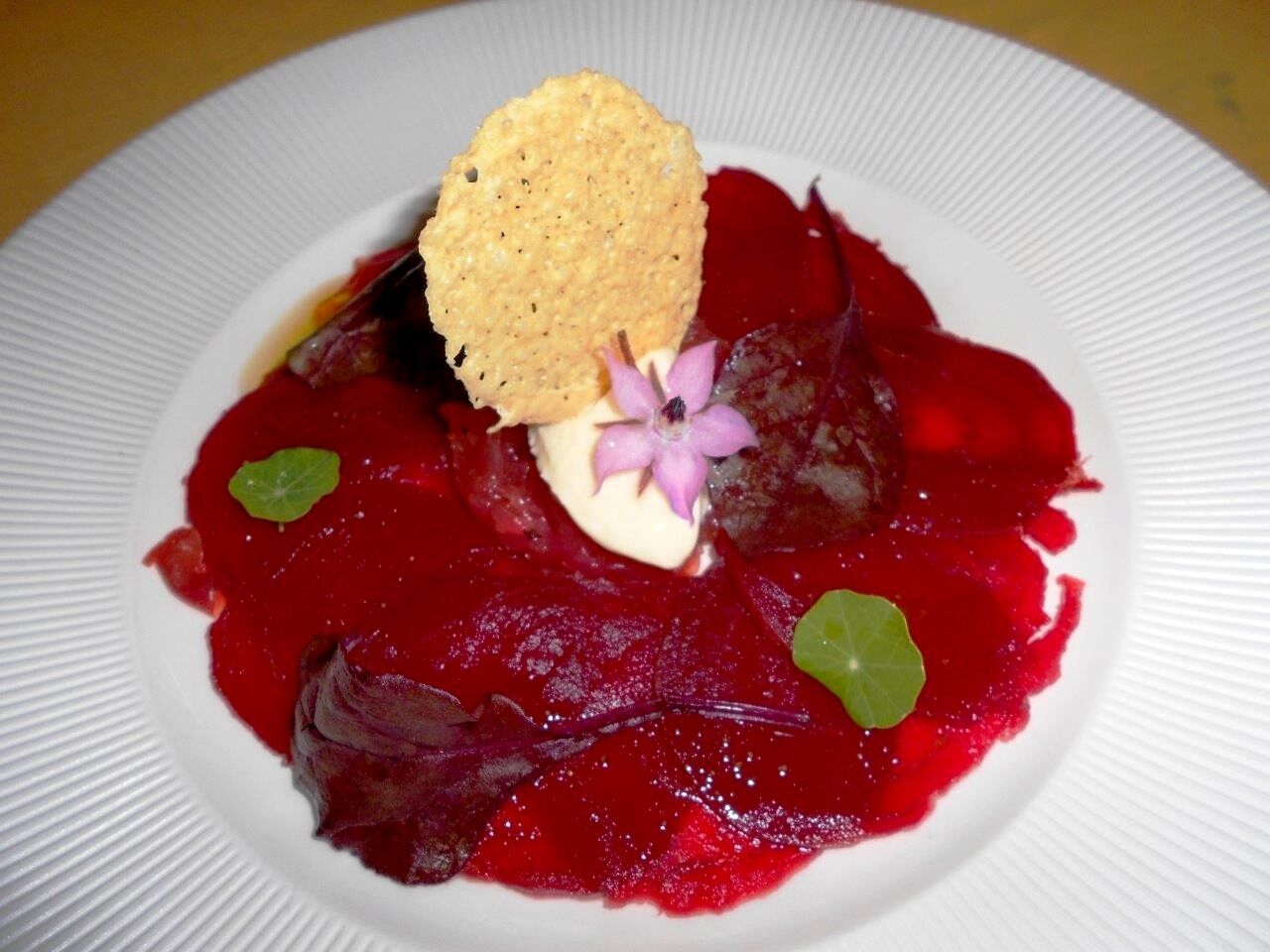 Une assiette blanche contenant un plat à base de betterave, agrémenté d’une fleur et d’un biscuit salé.