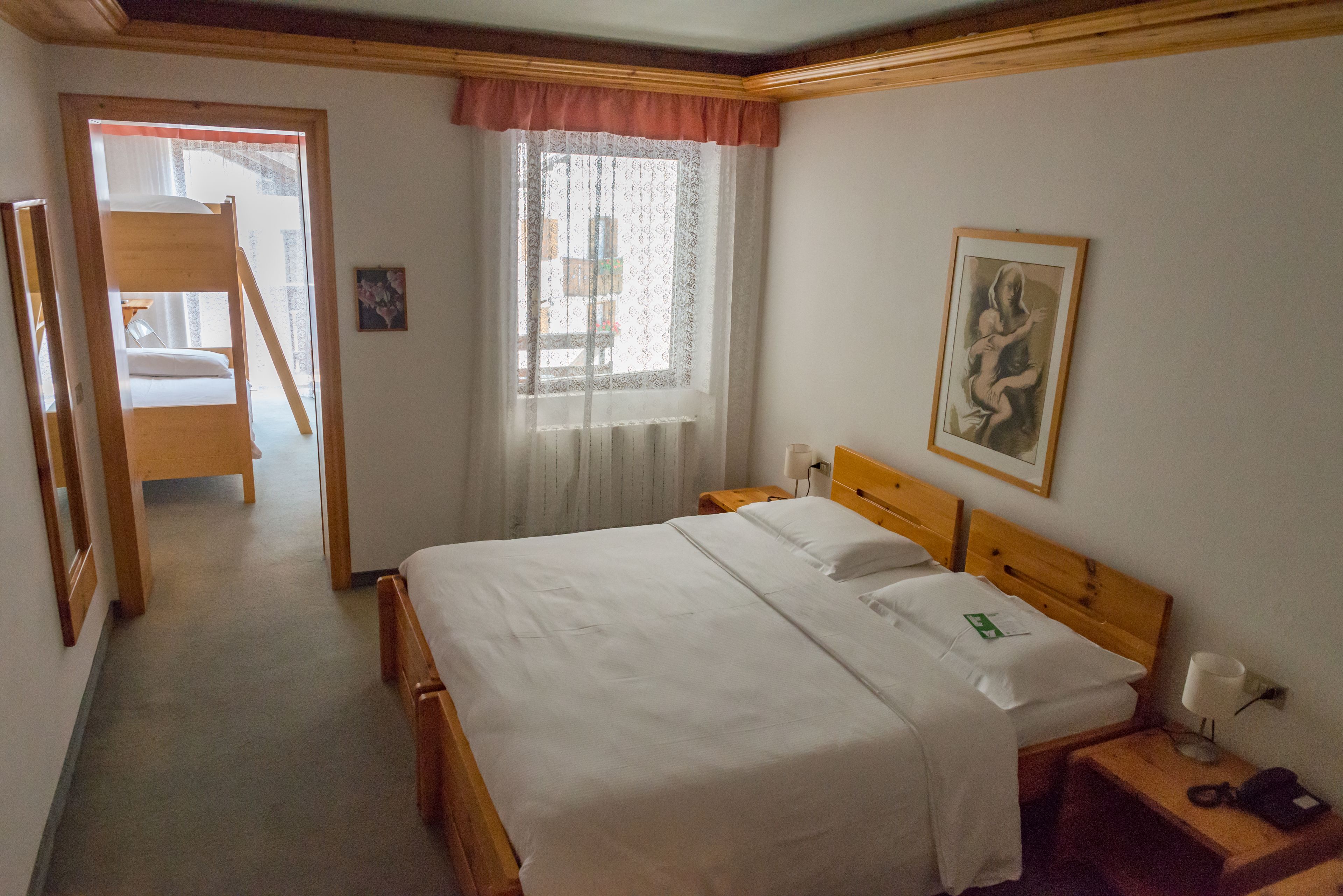 A bedroom with a wooden bed, a nightstand, a lamp, a framed picture on the wall, and a window with sheer curtains.