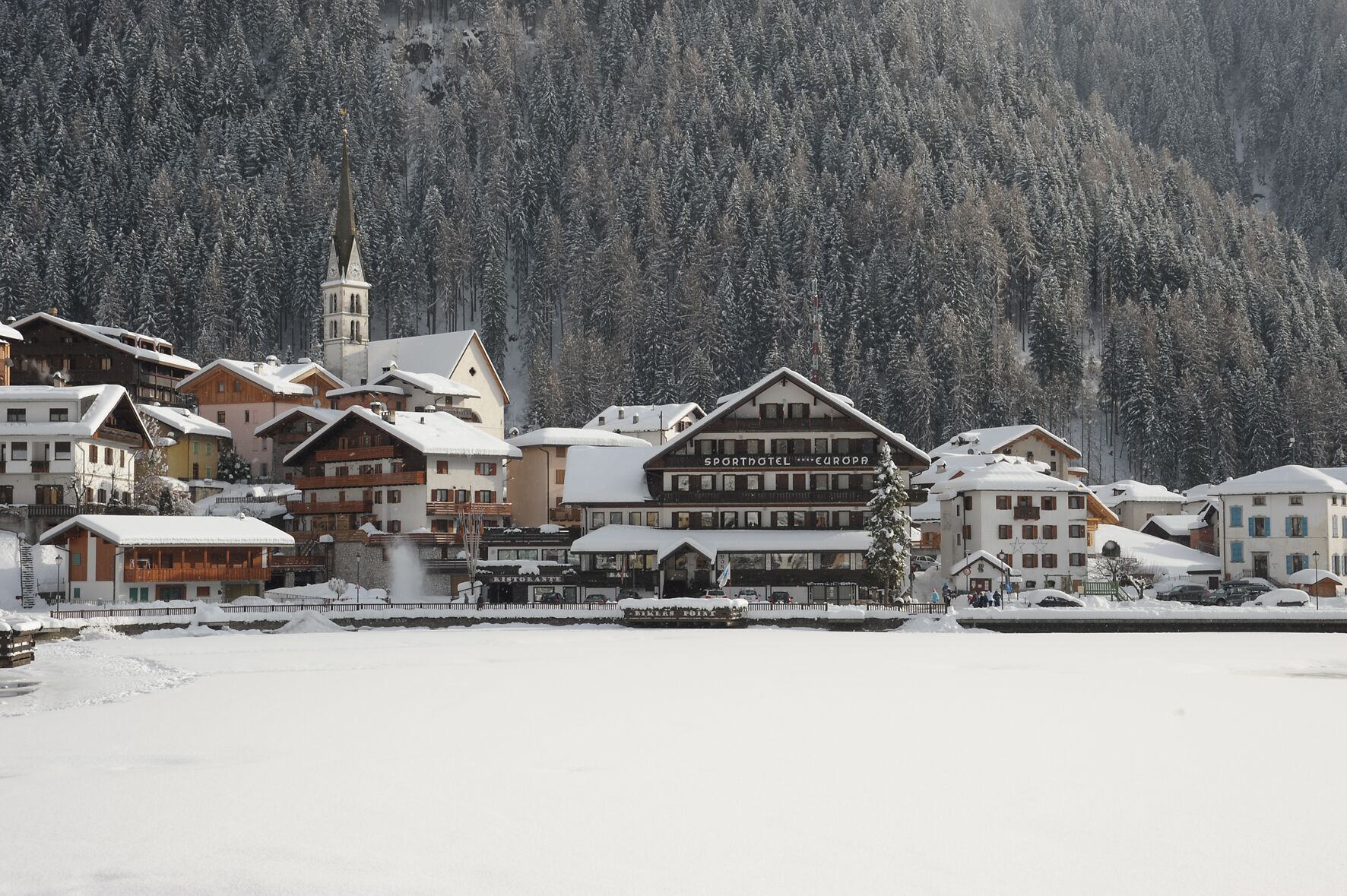 A snow-covered town with traditional Alpine architecture, including a prominent hotel with a sign reading "SPORTHOTEL EUROPA."