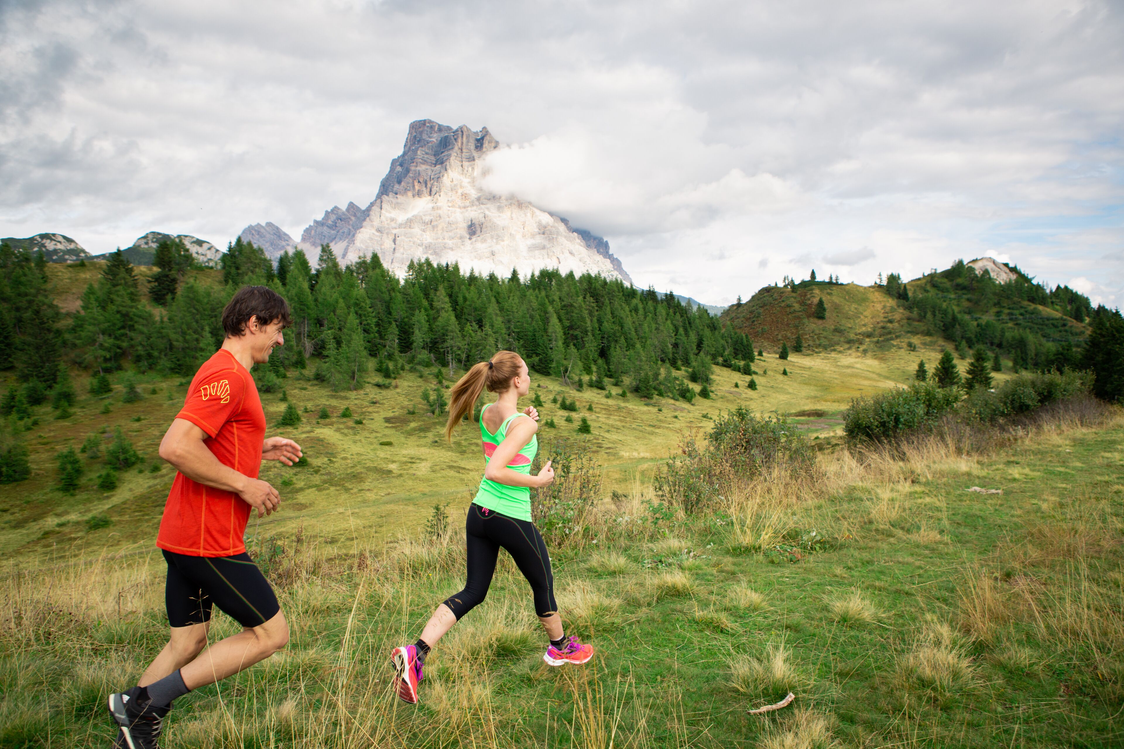 Two people running on a grassy field with mountains in the background.