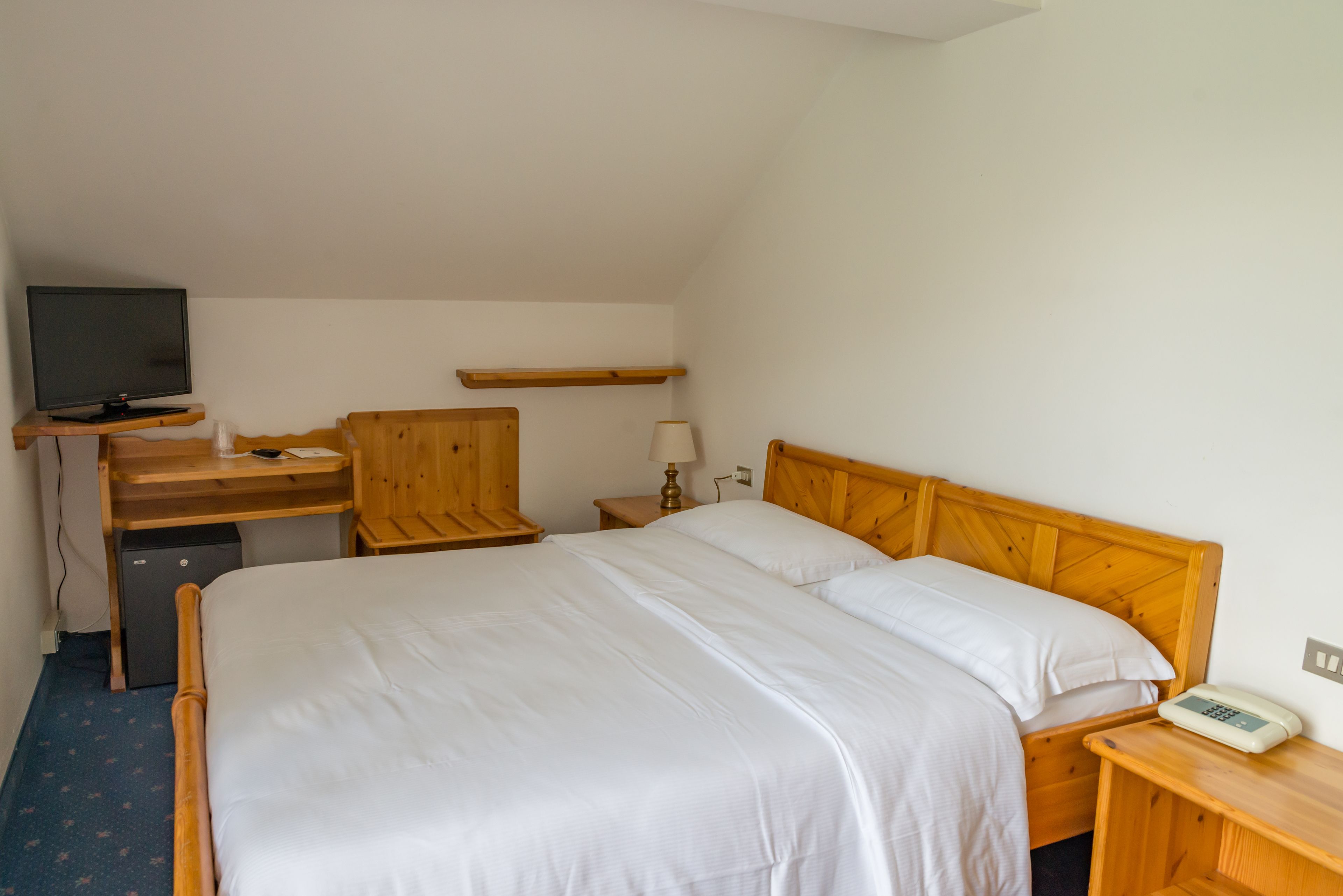A bedroom with a bed, a desk with a television, a lamp, and a bedside table with a phone.