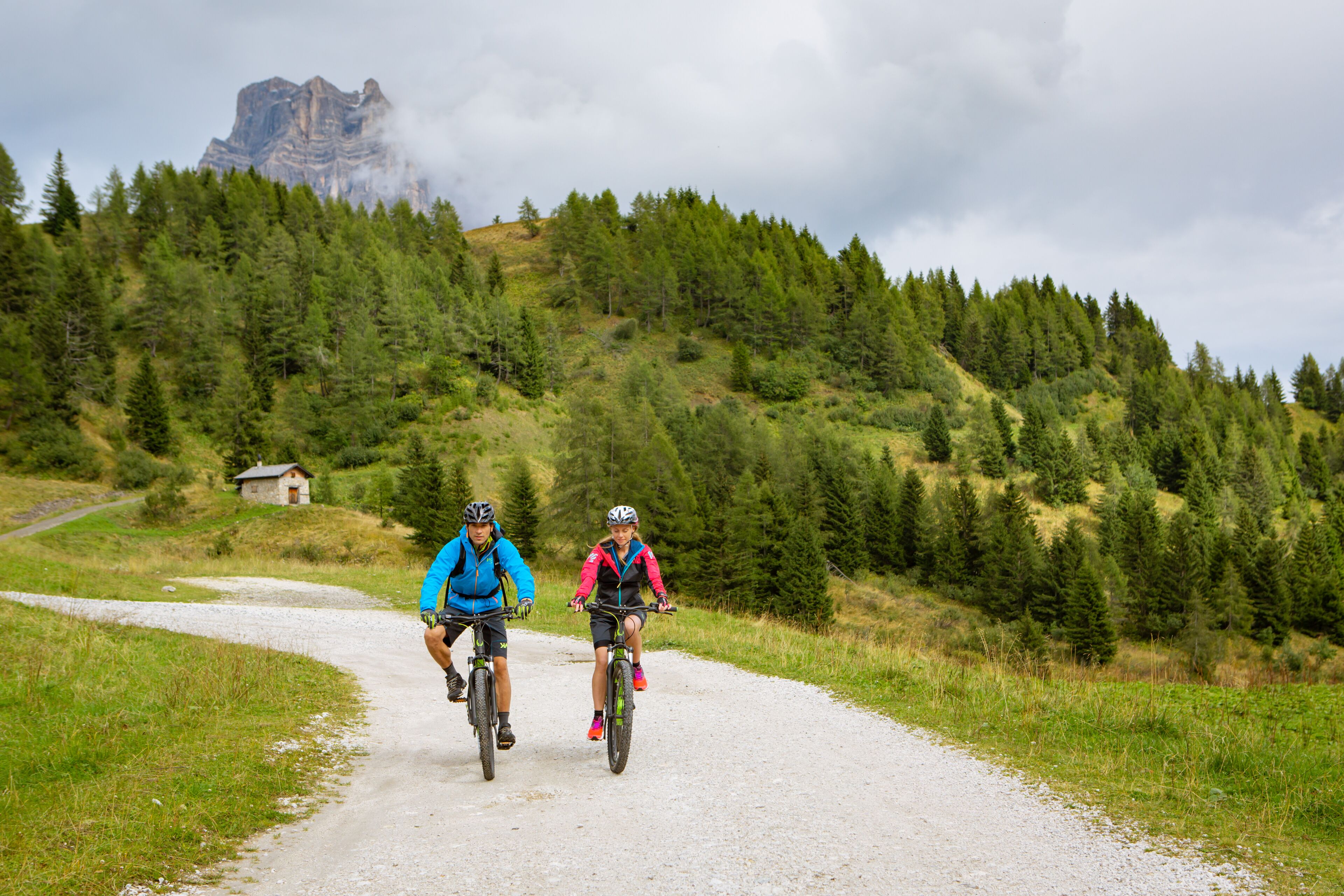 Two cyclists on a gravel path surrounded by dense forest and a mountain in the background.