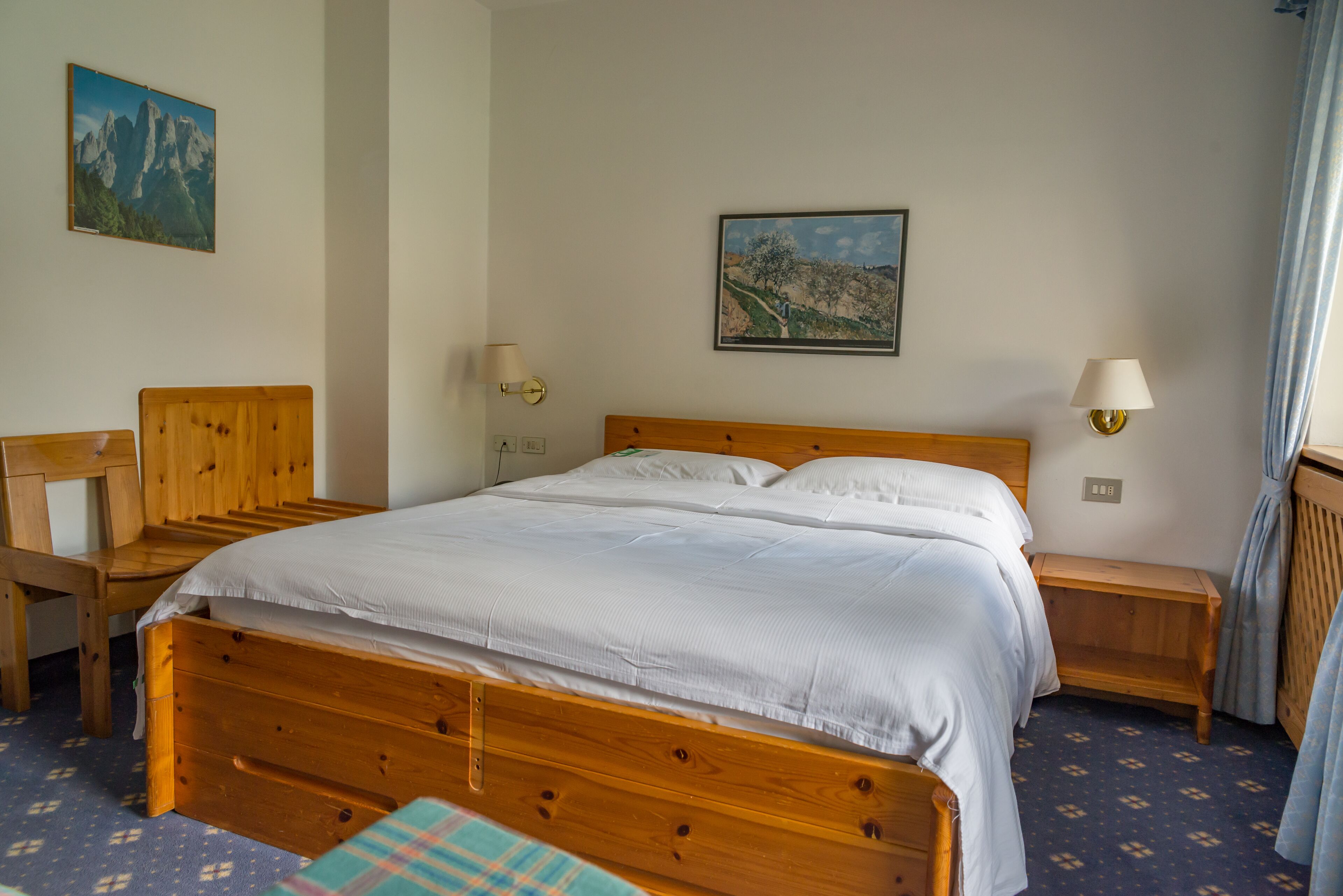 A neatly made wooden bed with white linens, a wooden nightstand with a lamp, a chair, and a small table.