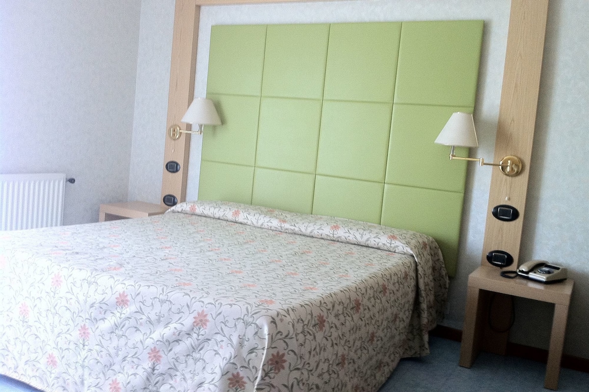 A hotel room with a bed, two bedside lamps, a nightstand with a phone, and a radiator.
