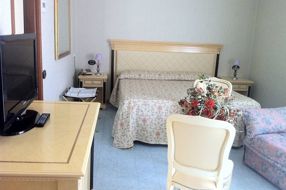 A hotel room with a bed, a desk with a television, a chair, and a bedside table with a lamp.