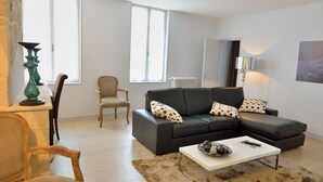A modern living room with a black sectional sofa, a white coffee table, and a beige armchair. There is a large window providing natural light, a radiator, and a lamp on a side table.