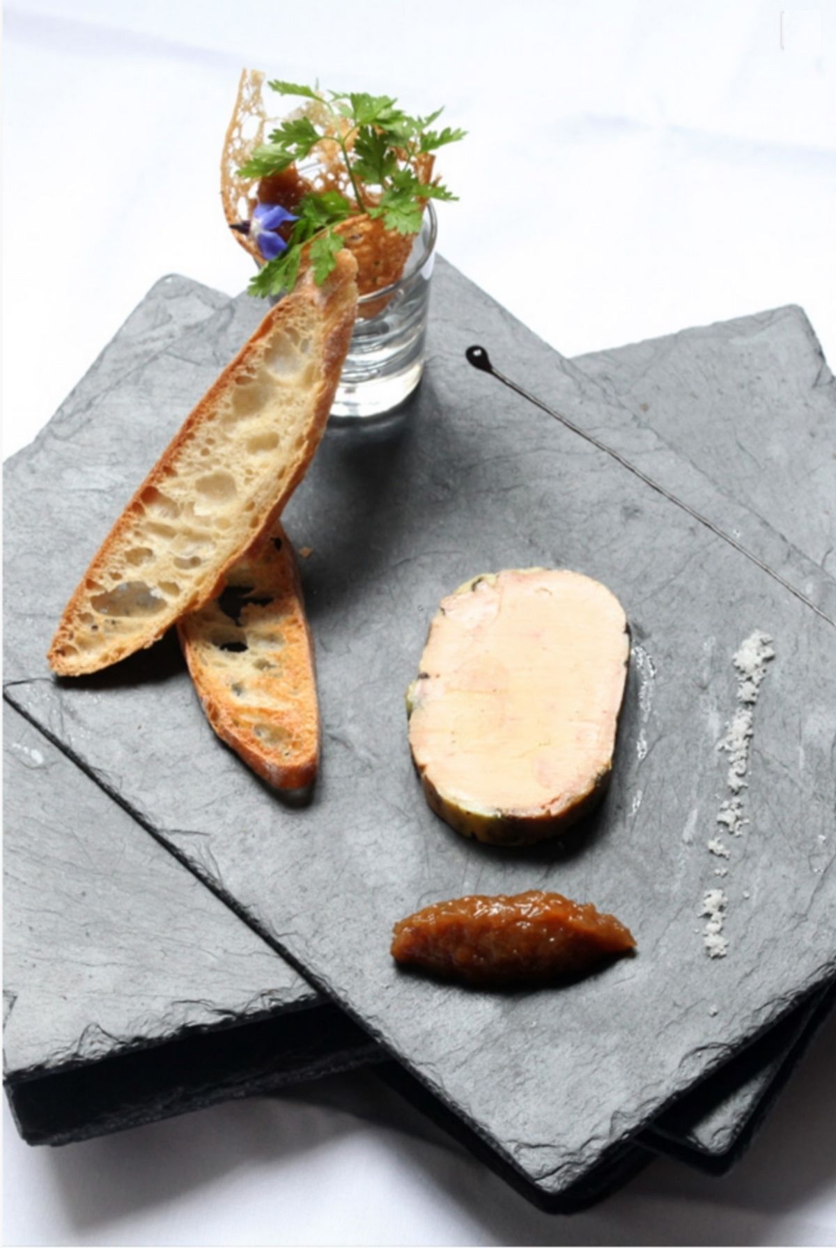 A slate serving board with a piece of bread, a slice of meat, and a small jar of sauce.