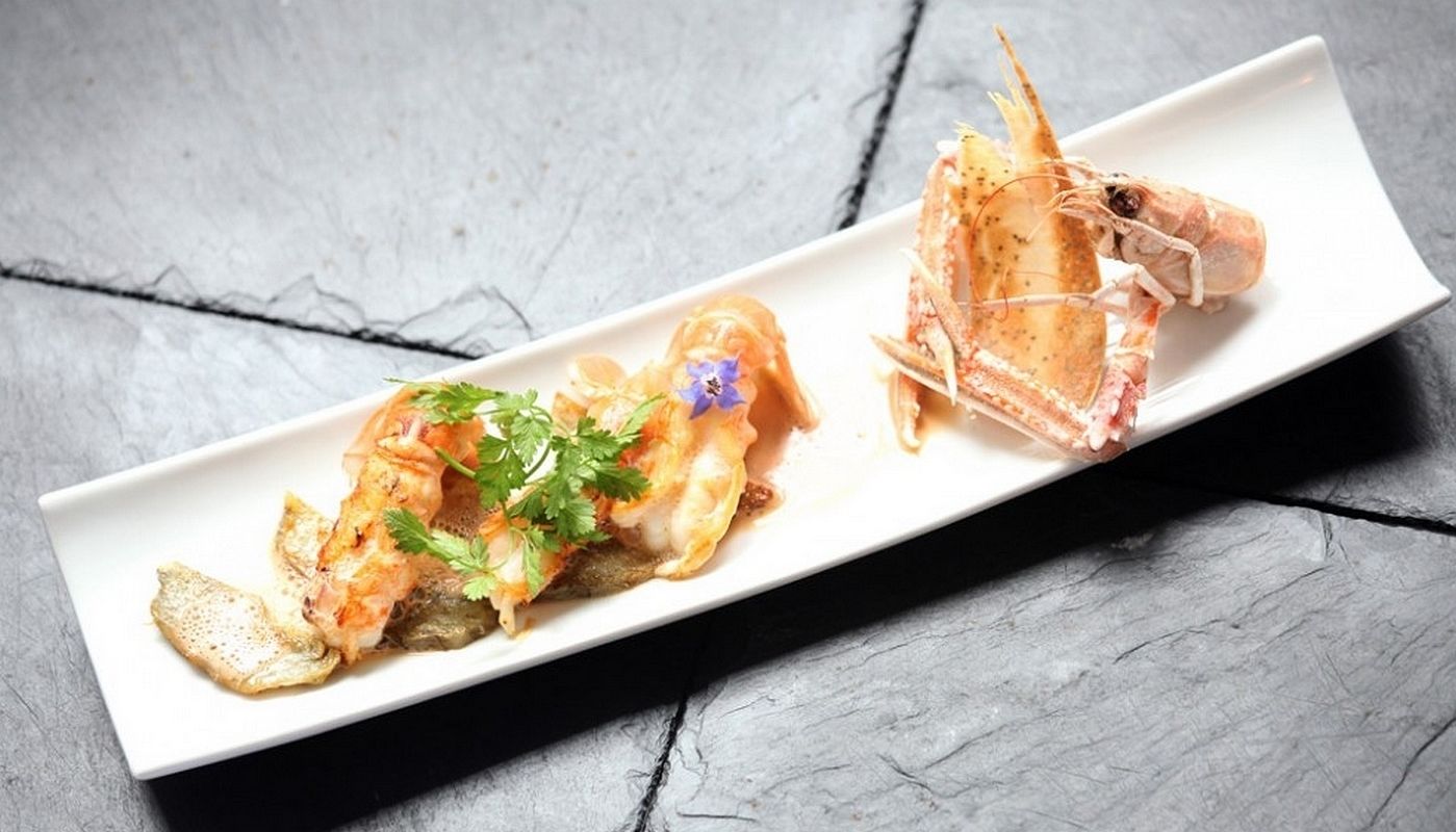 A rectangular white plate with two seafood dishes, one with a fish and shrimp garnish and the other with a crab, on a textured gray surface.