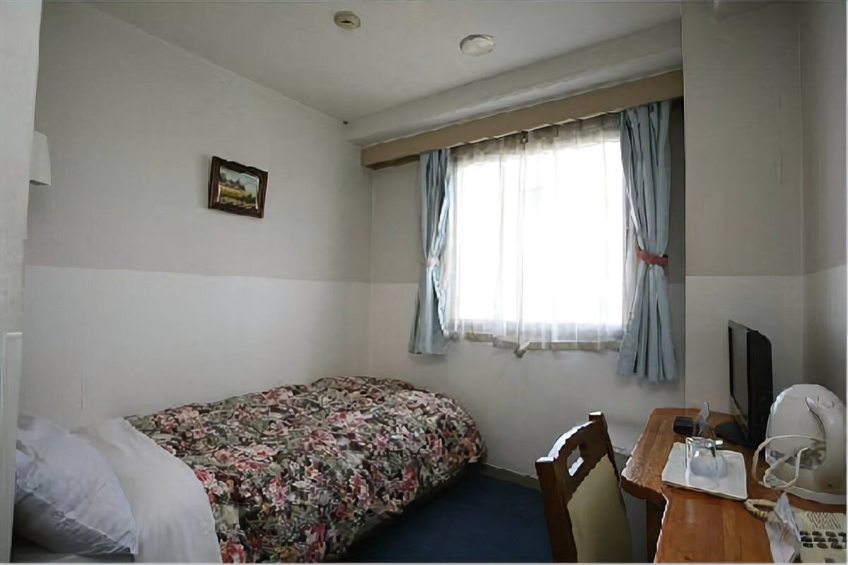 Ein kleiner, schlichter Raum mit einem Einzelbett, einem Schreibtisch mit Stuhl, einem Fernseher und einem Fenster mit Vorhängen.