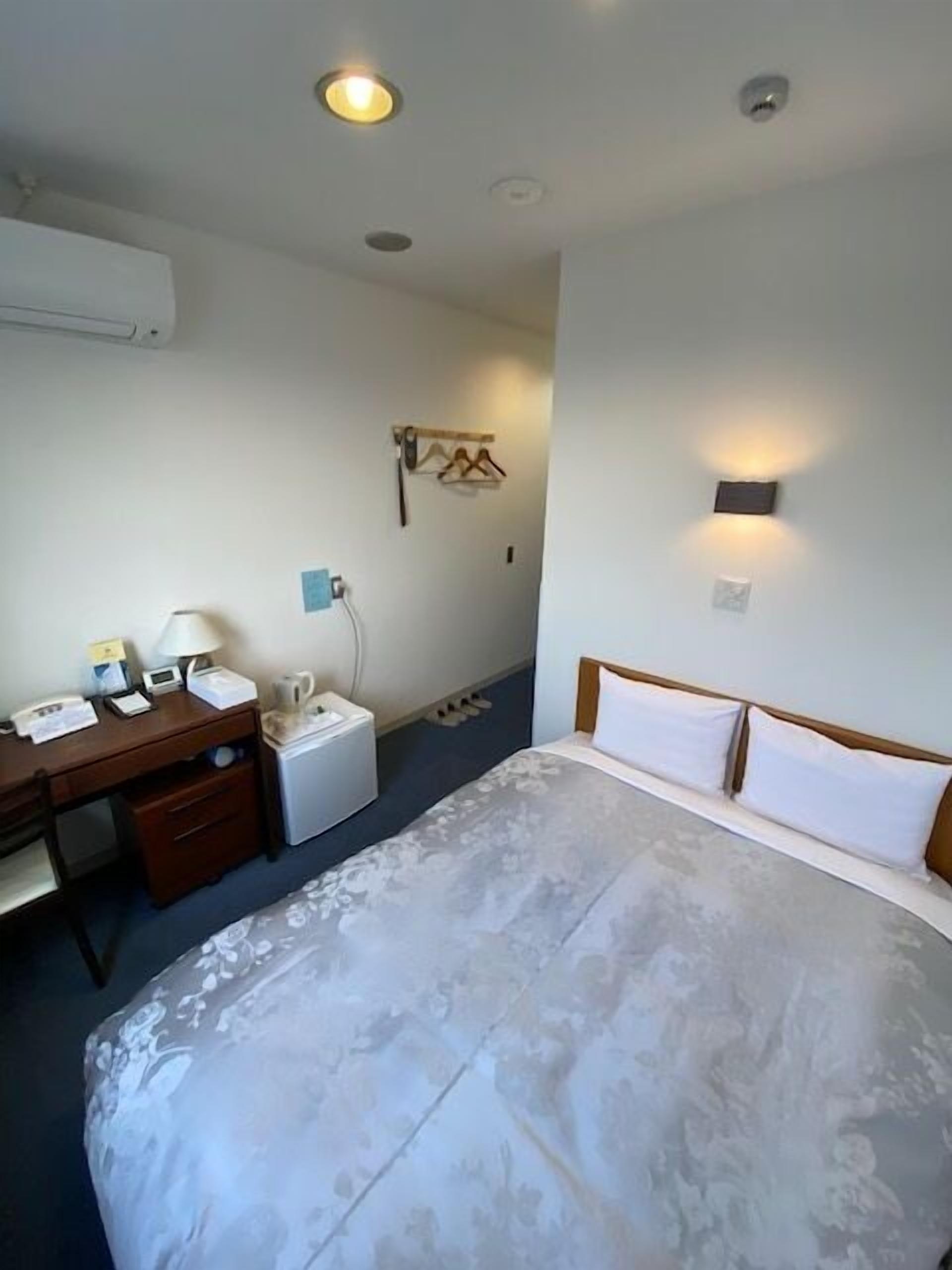 A hotel room with a bed, a desk, a chair, a small table, a wall-mounted heater, and a wall-mounted light fixture.