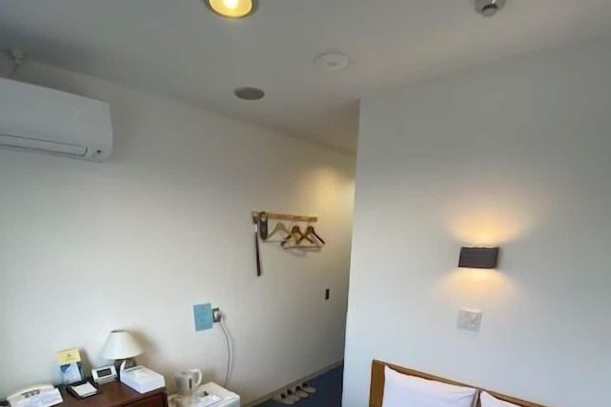 A hotel room with a bed, a desk, a chair, a small table, a wall-mounted heater, and a wall-mounted light fixture.