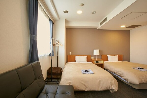 A hotel room with two beds, a sofa, a bedside table, a lamp, and a hanging coat rack.