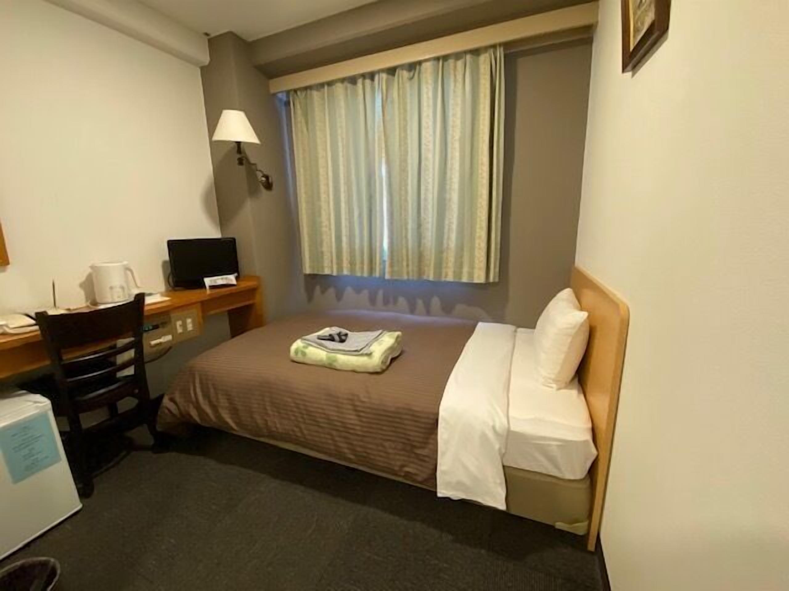 A hotel room with a bed, a desk with a computer, a chair, a lamp, and a window with curtains.