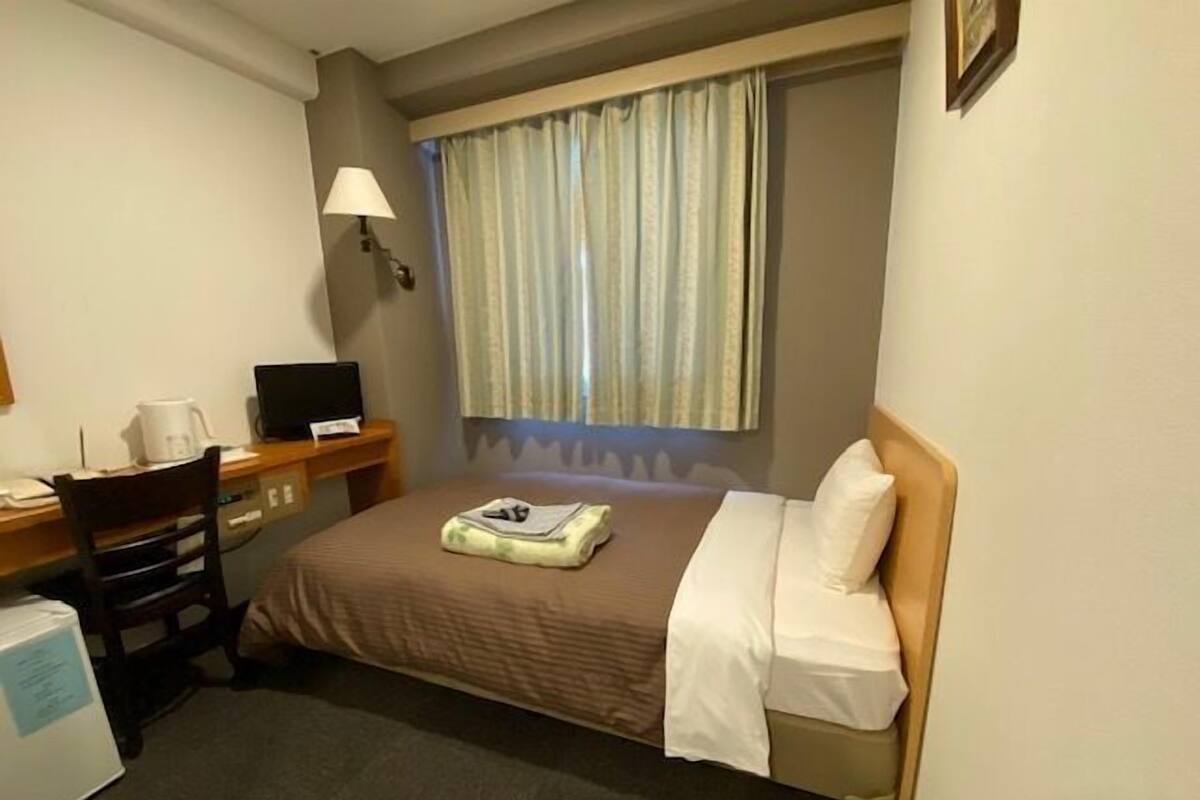 Ein Hotelzimmer mit einem Bett, einem Schreibtisch mit Computer, einem Sessel und einem Fenster mit Vorhängen.