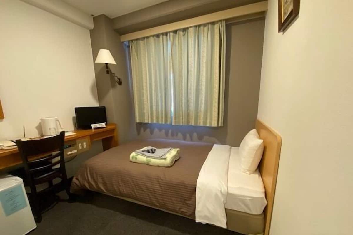 A hotel room with a bed, a desk with a computer, a chair, a lamp, and a window with curtains.