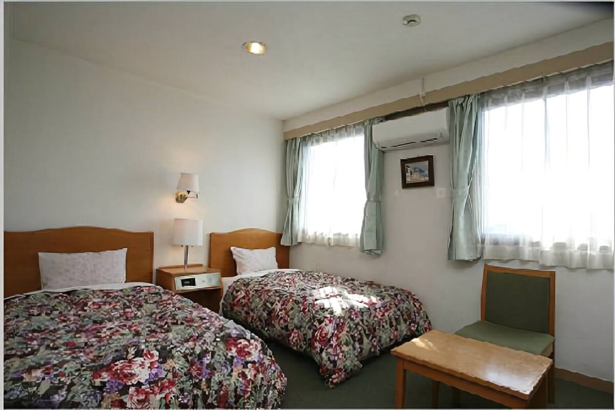 Ein Hotelzimmer mit zwei Betten, einem Nachttisch mit Lampe, einem Sessel und einem Fenster mit Vorhängen.