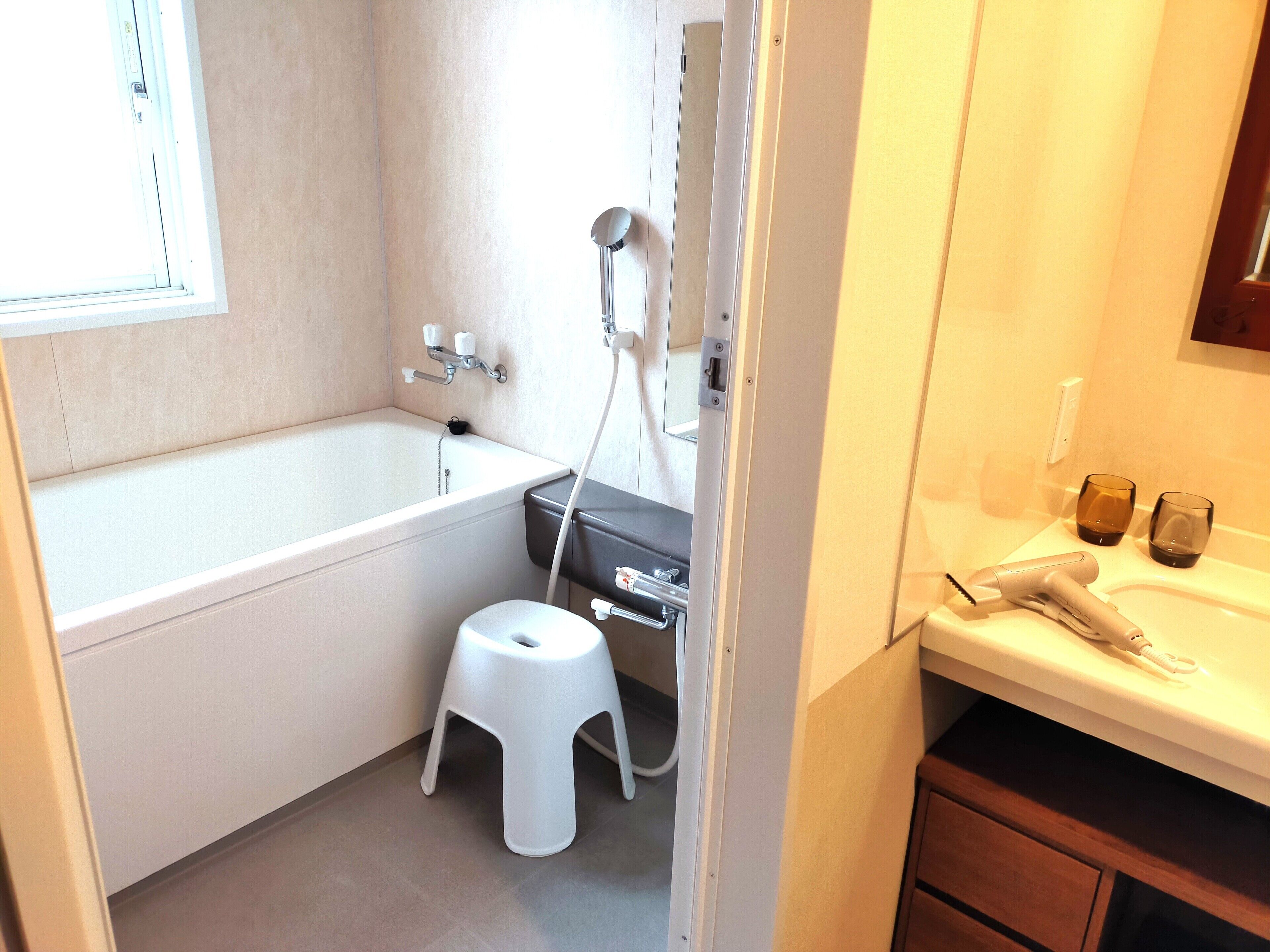 A bathroom with a bathtub, a toilet, a small white stool, a hairdryer, and two glasses on the counter.
