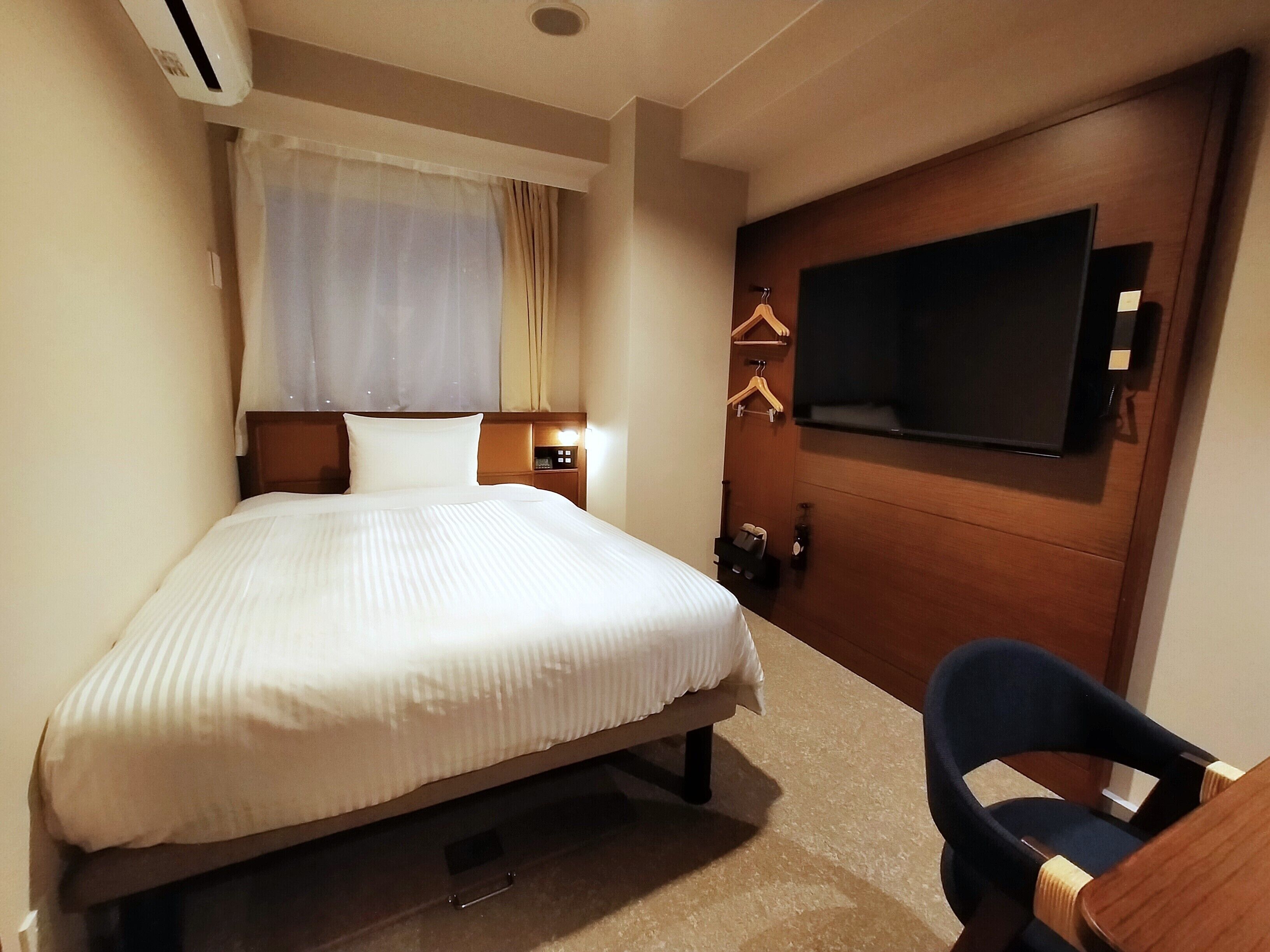 Une chambre d’hôtel comprenant un lit, un téléviseur fixé au mur, un bureau avec une chaise et une fenêtre avec des rideaux.