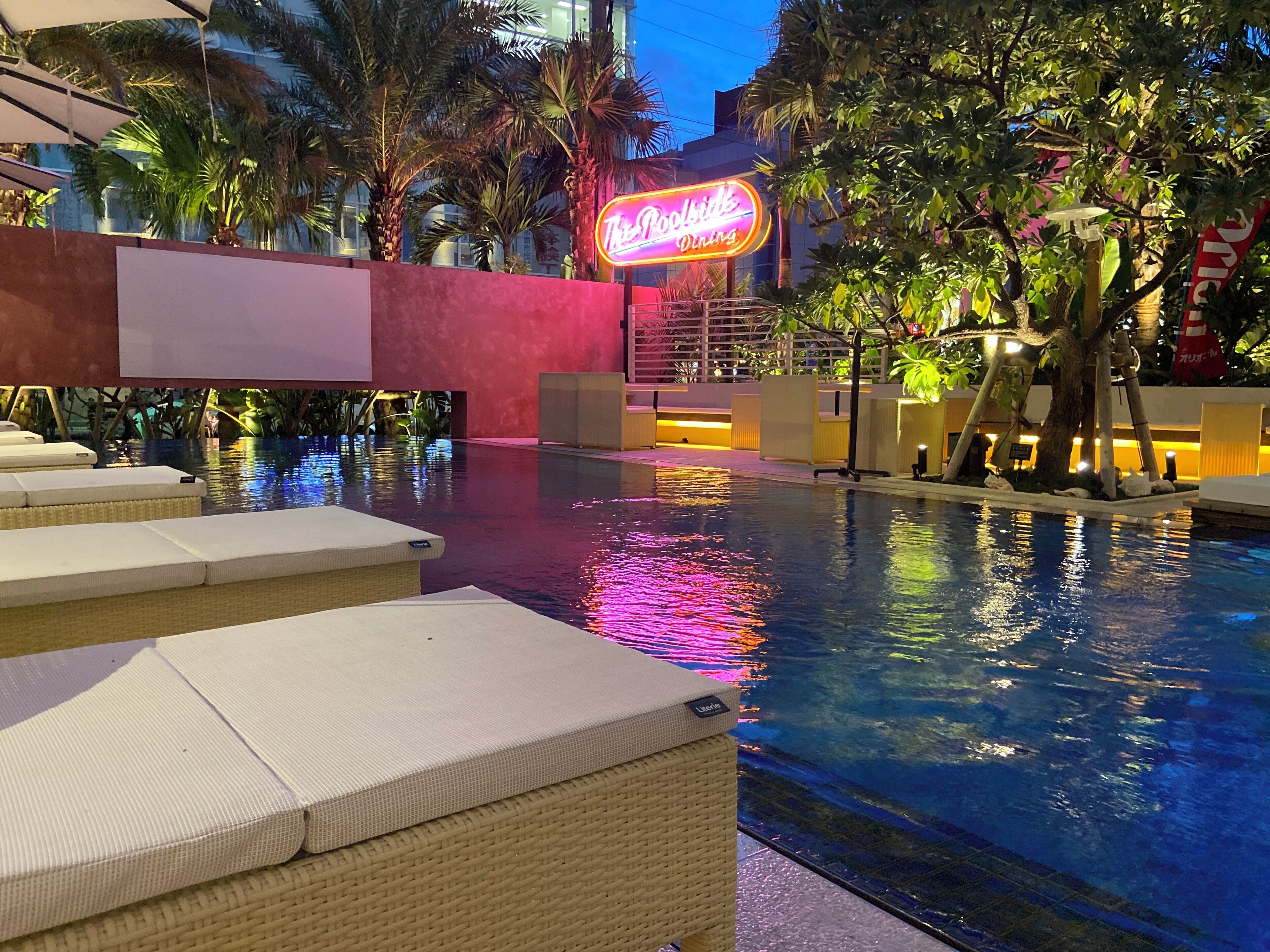 A poolside area with lounge chairs, a neon sign for 'Poolside Dining', and palm trees.