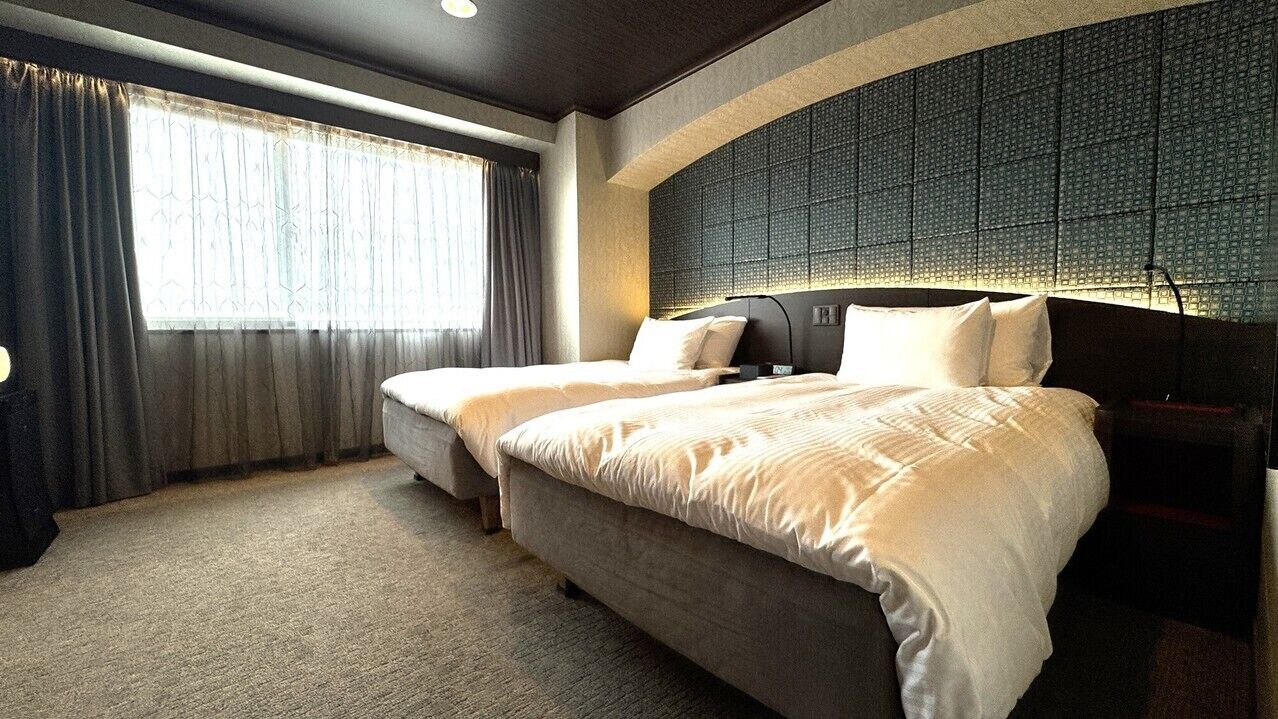 A hotel room with two beds, a large window with curtains, and a textured wall behind the beds.