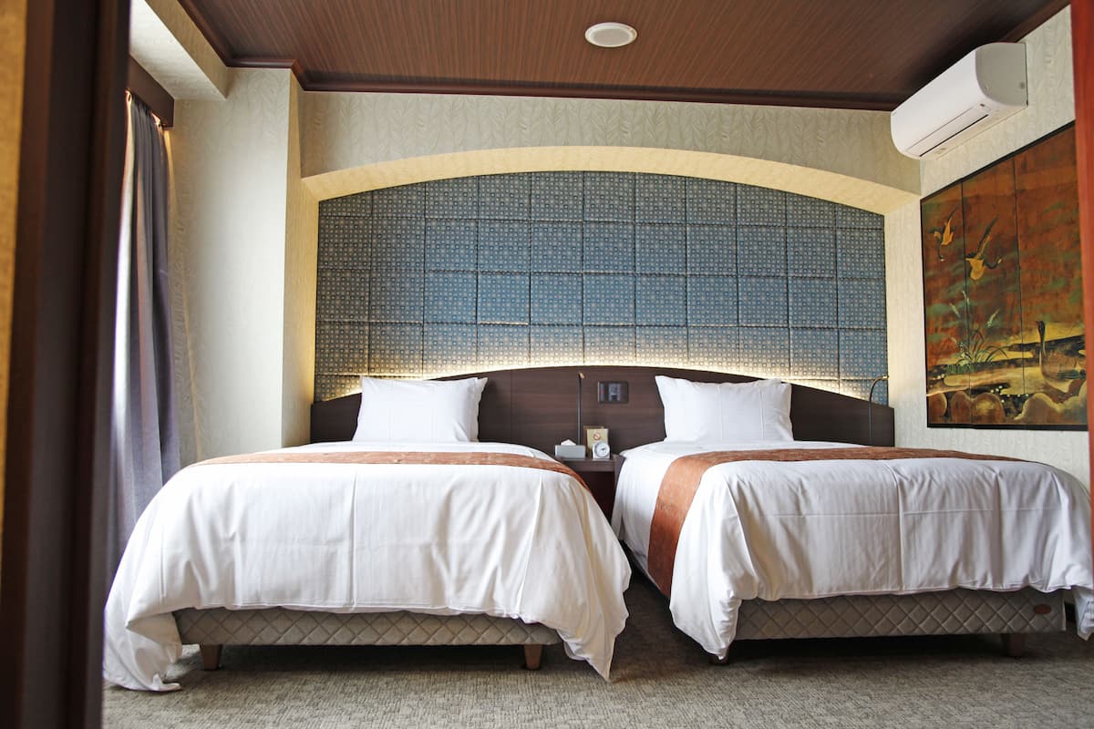 A hotel room with two beds, a large decorative headboard, a painting on the wall, and an air conditioning unit.