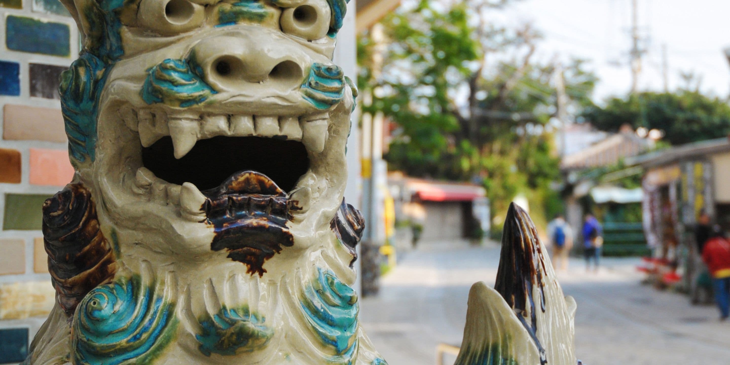 A close-up of a decorative lion head sculpture with a wide mouth and teeth, set against a backdrop of a street with buildings and people.